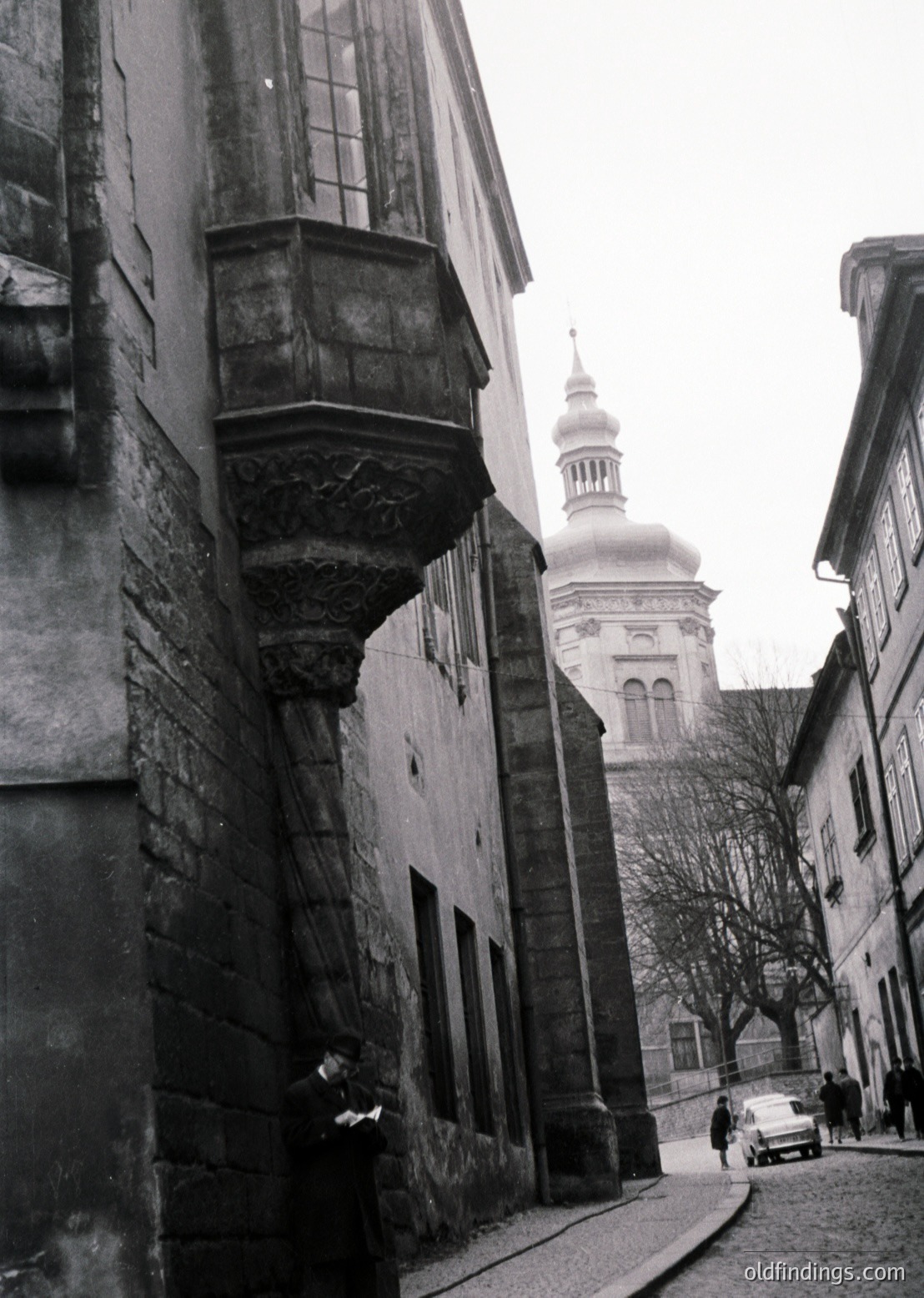 Narrow cobblestone street in a historic European town, flanked by stone buildings with ornate balconies. A vintage car and pedestrians add life to the scene. Dominating the background is a distinctive white-domed church with arched windows, likely Baroque or Renaissance Revival.