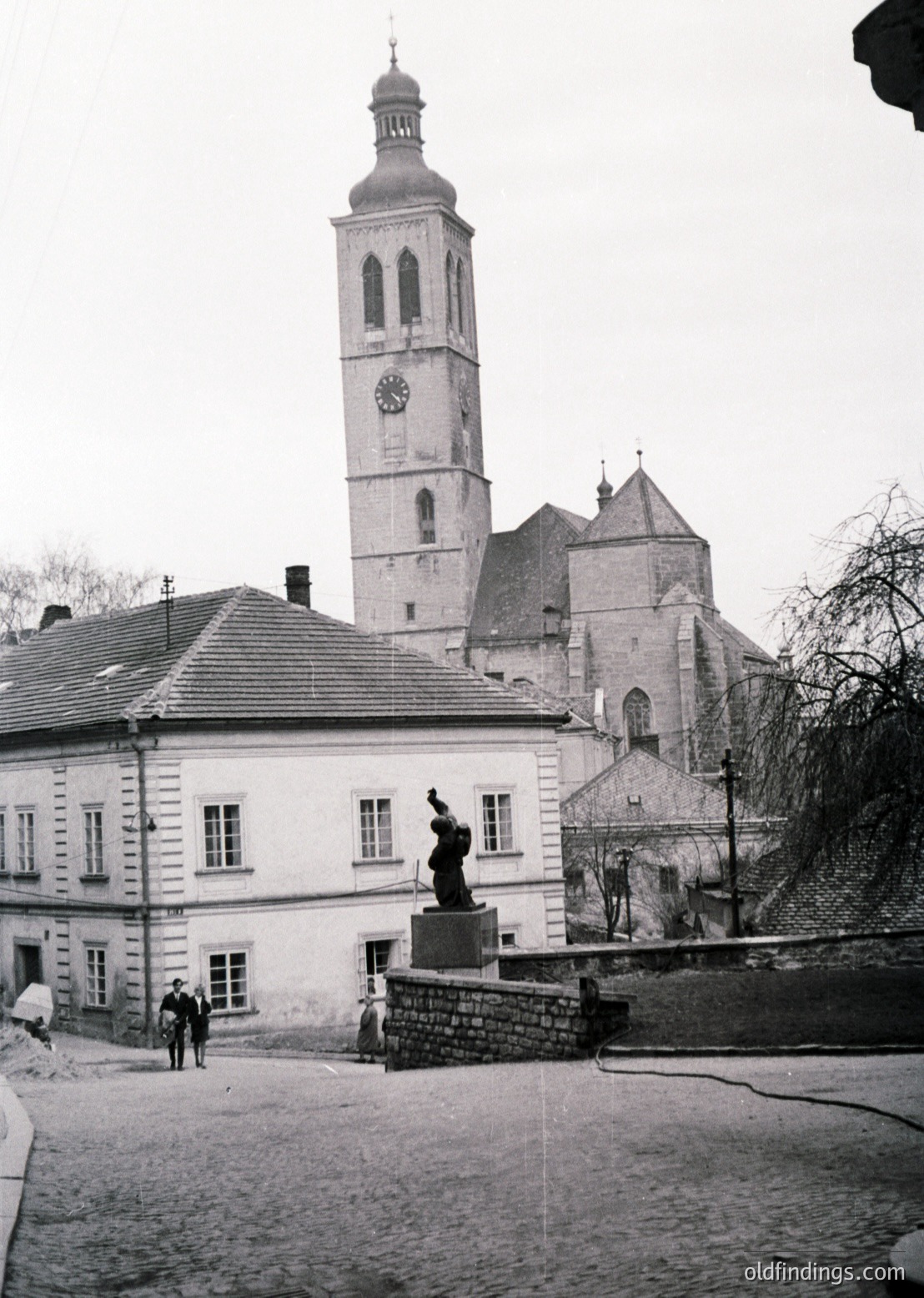 Historic European town square featuring a prominent **clock tower** with Gothic Revival architecture, likely from the **19th century**. Foreground statue depicts a **musician** atop a stone pedestal. Residential buildings with **slate roofs** and **whitewashed walls** frame the scene. Overcast sky enhances the vintage atmosphere.