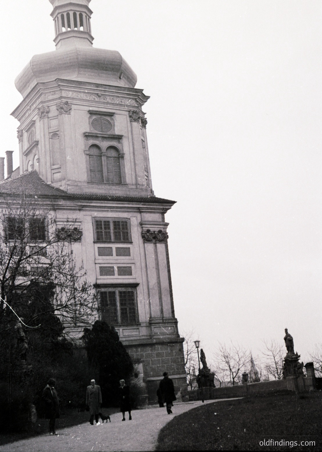 Neoclassical stone tower with domed roof and symmetrical windows, flanked by bare trees and a statue pedestal. Mid-20th century urban scene with pedestrians in winter attire.