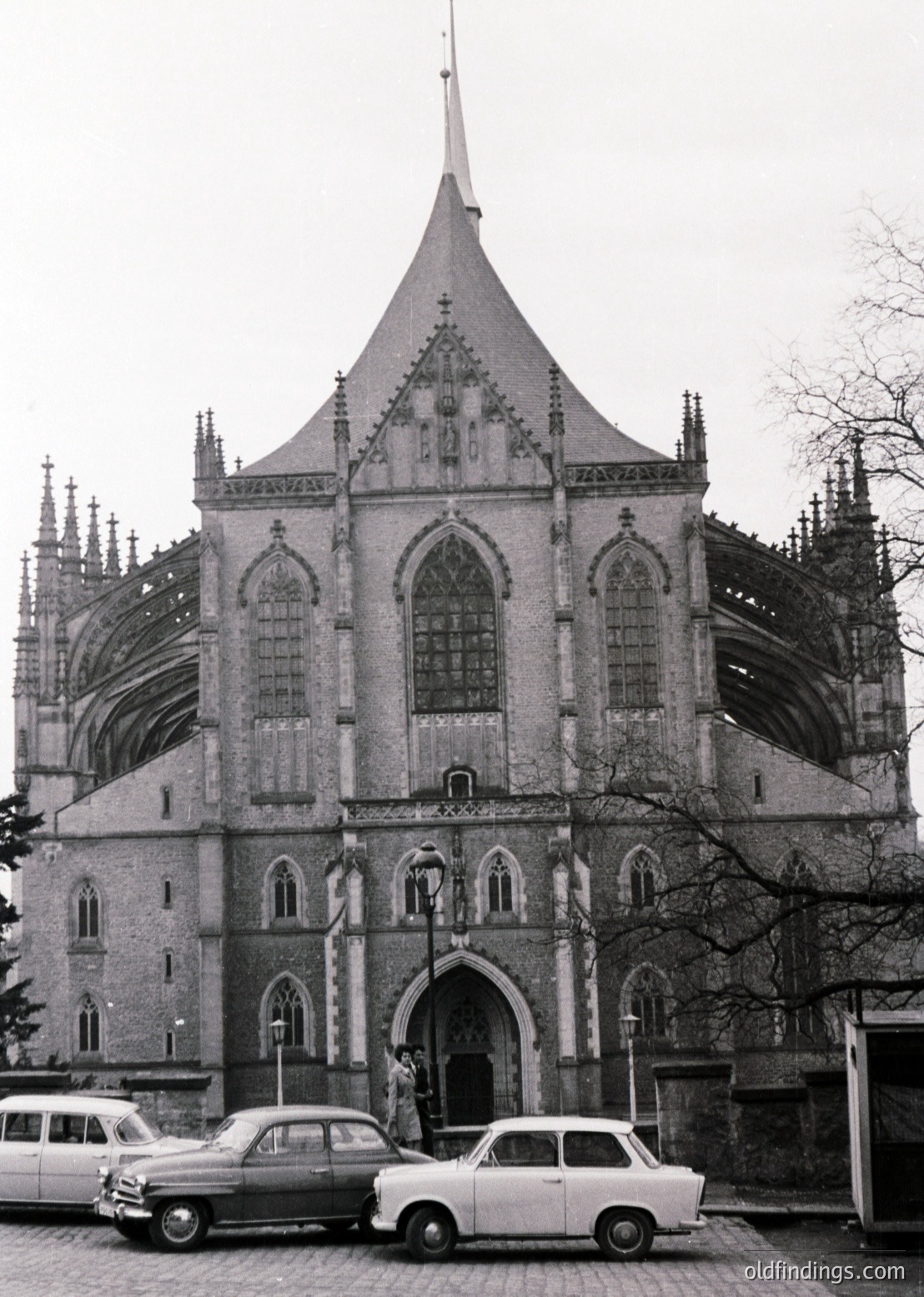 Gothic Revival church façade with pointed arches, ribbed vaulting, and a central spire, likely from the 19th century. Two vintage cars (1950s–60s) parked in front, one with a chrome grille and tailfins. Bare trees suggest late autumn/winter. Architectural details include ornate stonework and stained-glass windows.