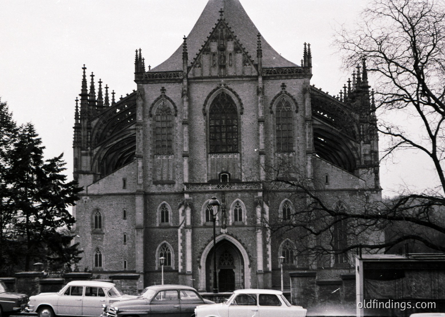 Gothic Revival church facade with pointed arches, spires, and intricate stonework, likely mid-20th century. Symmetrical design features a central entrance flanked by tall, narrow windows. Classic cars parked in front suggest historical urban setting.