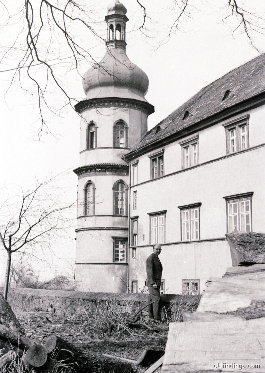 Neoclassical building with domed tower and arched windows, likely 19th-century European architecture. Man in dark suit poses on stone steps beside overgrown garden.