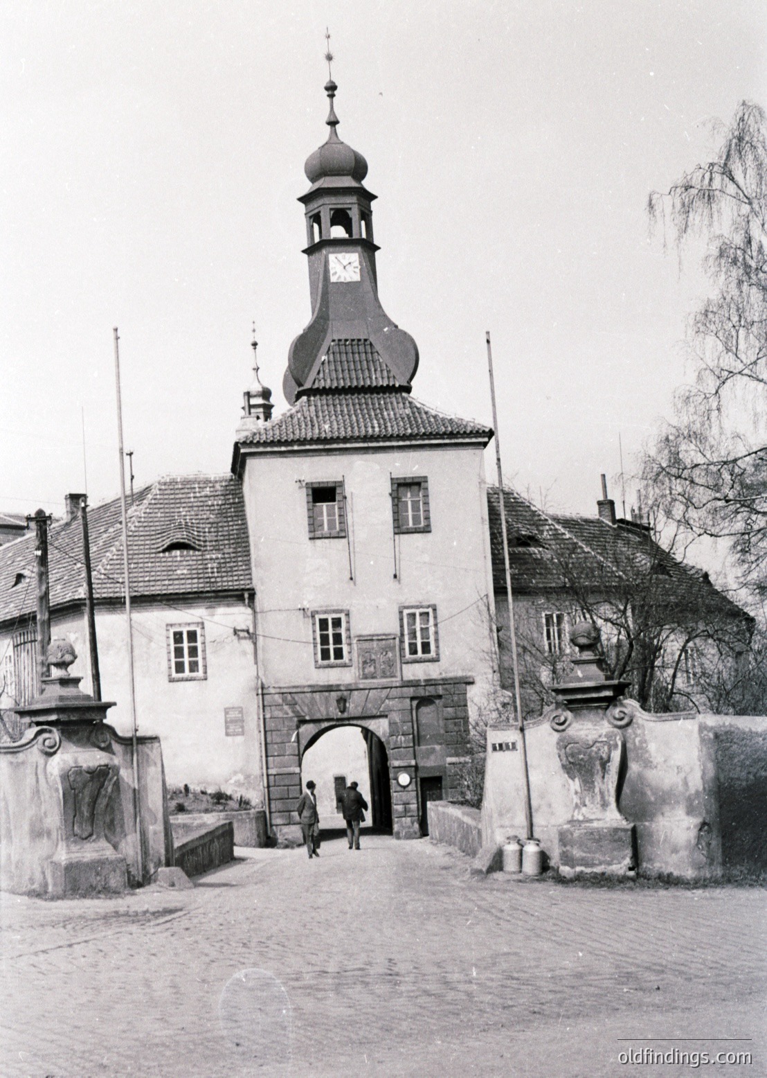 Historic town gate with Baroque-style tower and arched entrance, flanked by stone pillars. Cobblestone pathway leads through the gateway, with two figures walking. Architectural details include a clock on the tower and decorative stonework. Likely Eastern European, 18th–19th century.