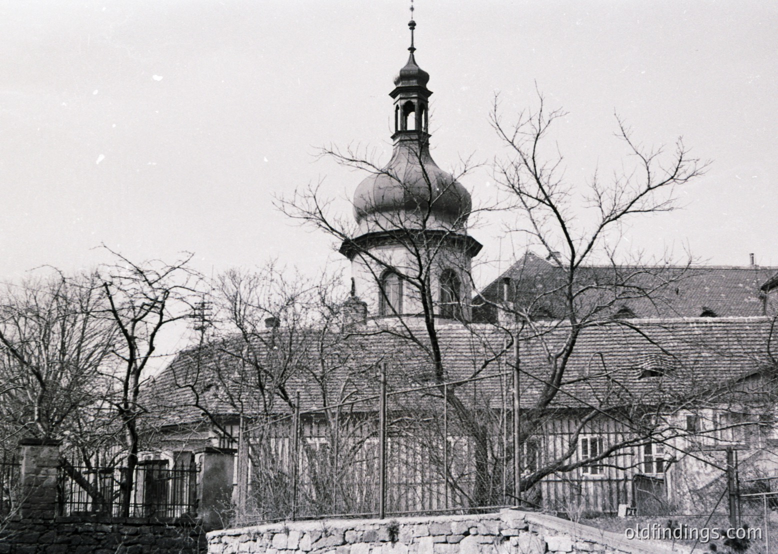 Black-and-white photograph of a domed church with a spire, framed by bare winter trees and a stone fence. Likely Eastern European architecture, mid-20th century.