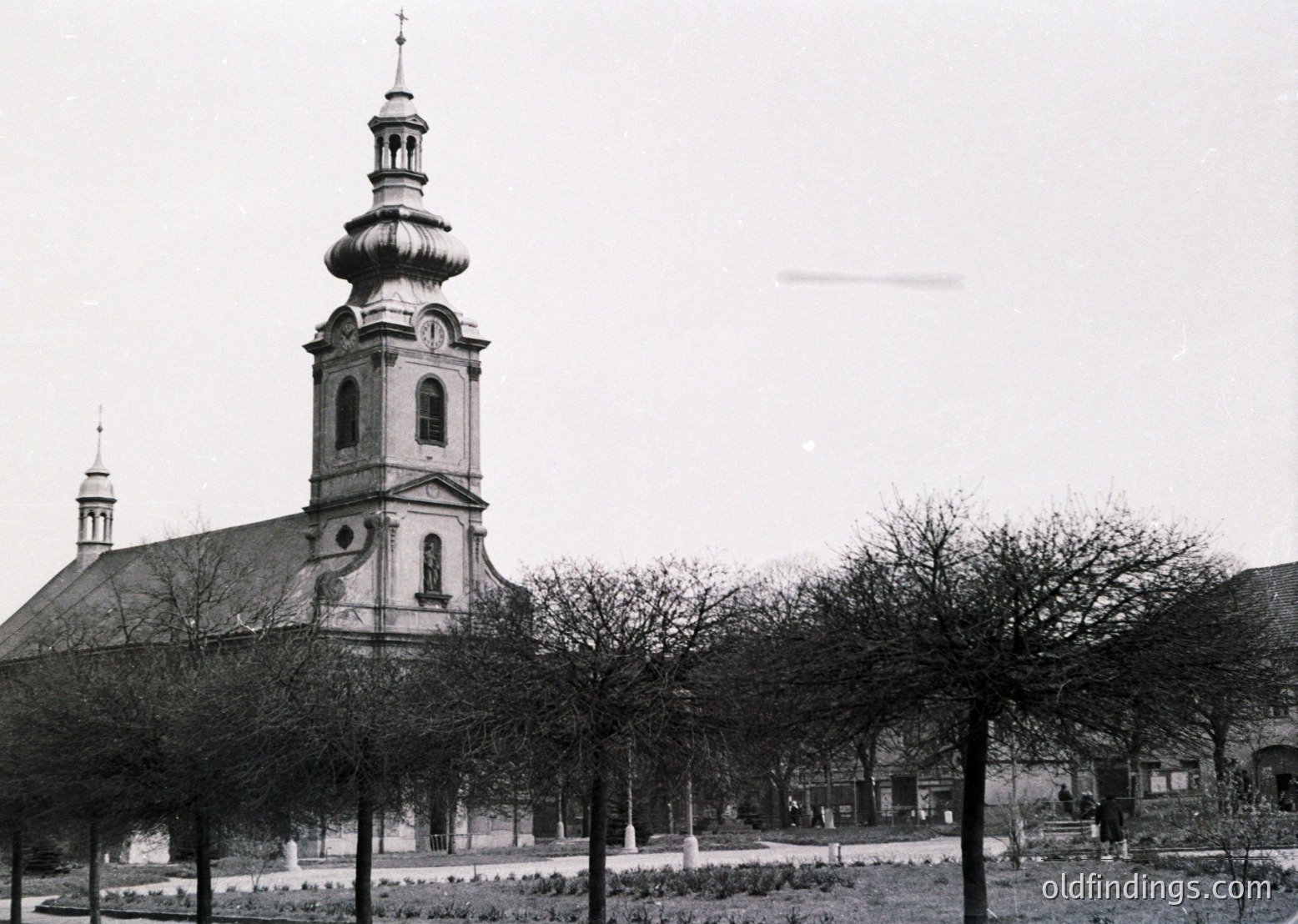 Neoclassical church with domed tower and symmetrical façade, flanked by trimmed trees in a courtyard. Mid-20th century European architectural style.