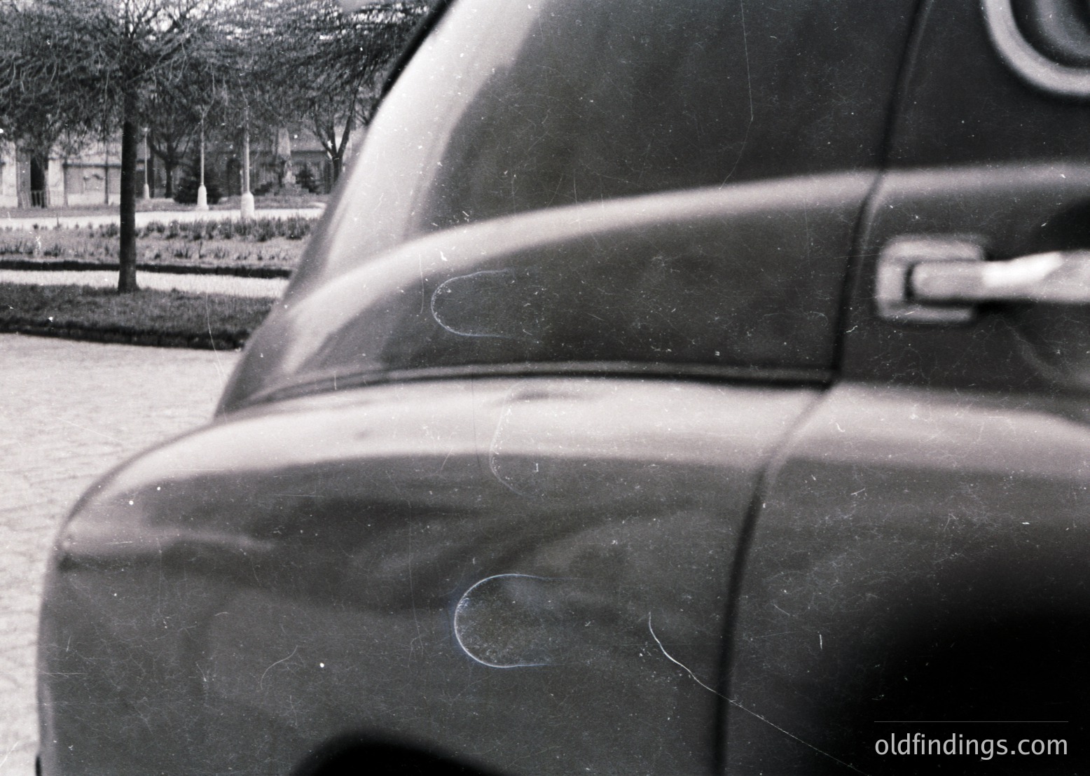 Close-up of a vintage car’s rear quarter panel, showing subtle scratches and minor paint imperfections. Blurred background reveals a landscaped park with trees and manicured hedges, suggesting an urban setting. Likely mid-20th century automotive design.