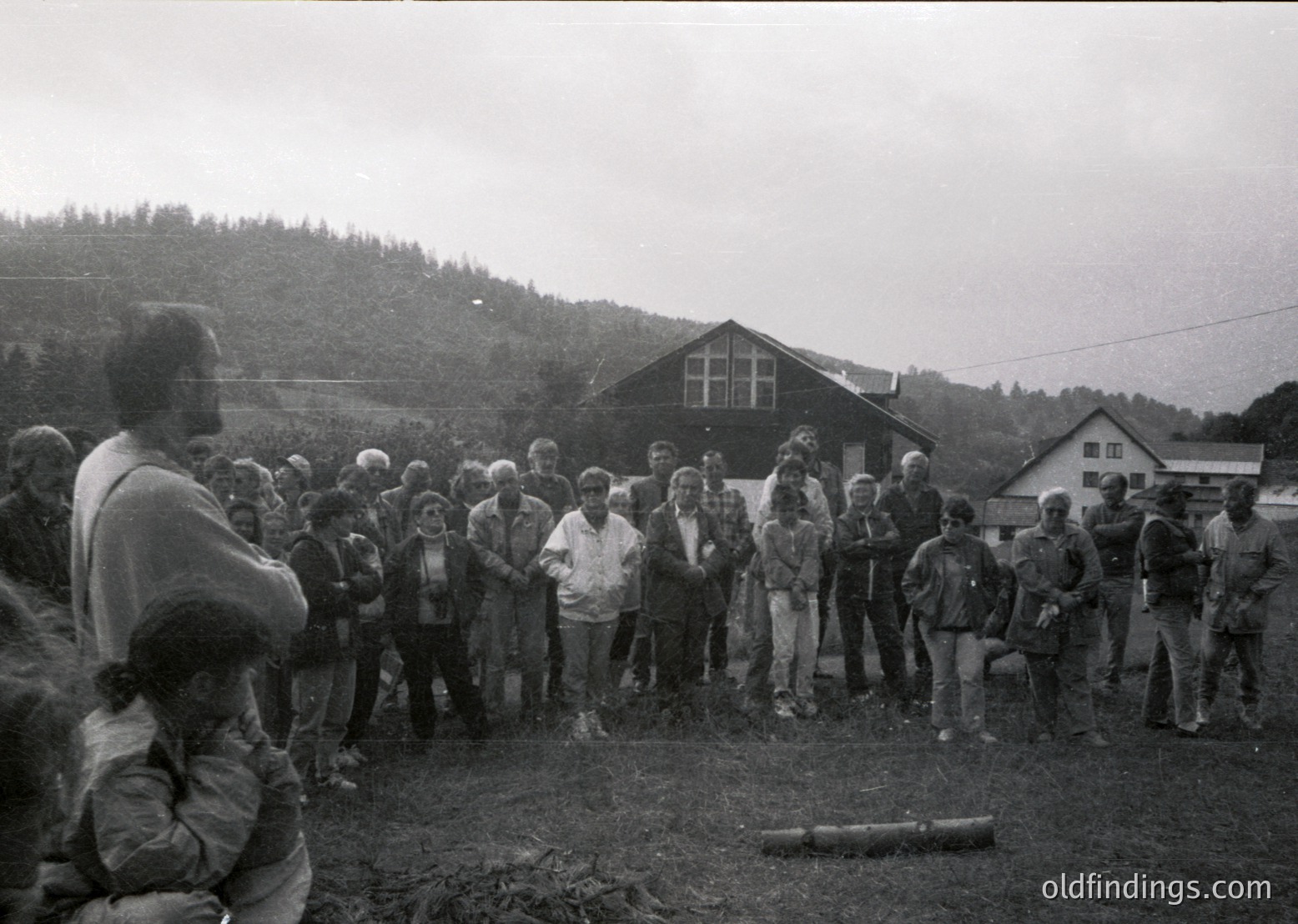 Black-and-white gathering of ~30 people in a rural outdoor setting, likely mid-20th century. Group stands on uneven terrain near a wooden building with gabled roof and a log stump in foreground. Attendees wear casual, utilitarian clothing—some in jackets, others in overalls—suggesting a communal or agricultural event. Overcast sky and forested hills in background.