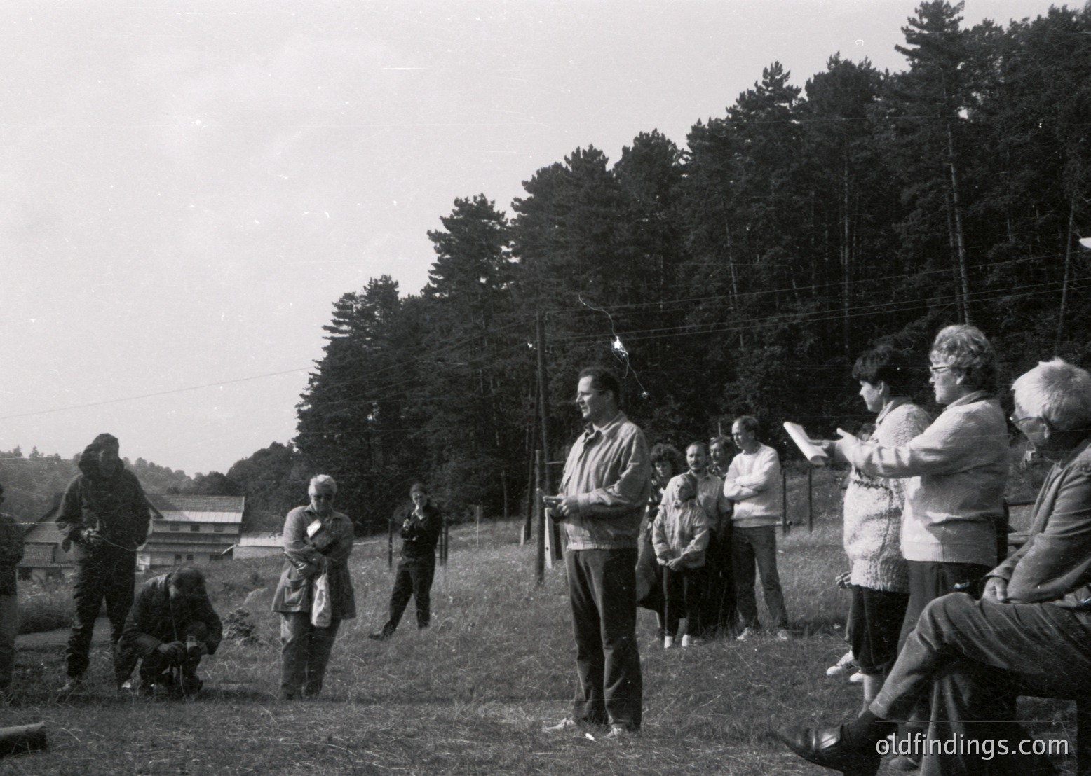 Black-and-white outdoor gathering in a rural setting, likely mid-20th century. A speaker addresses a seated audience of ~15 people in casual attire (cardigans, trousers, hats). Wooden fence and dense forest backdrop. Potential community event or public address.