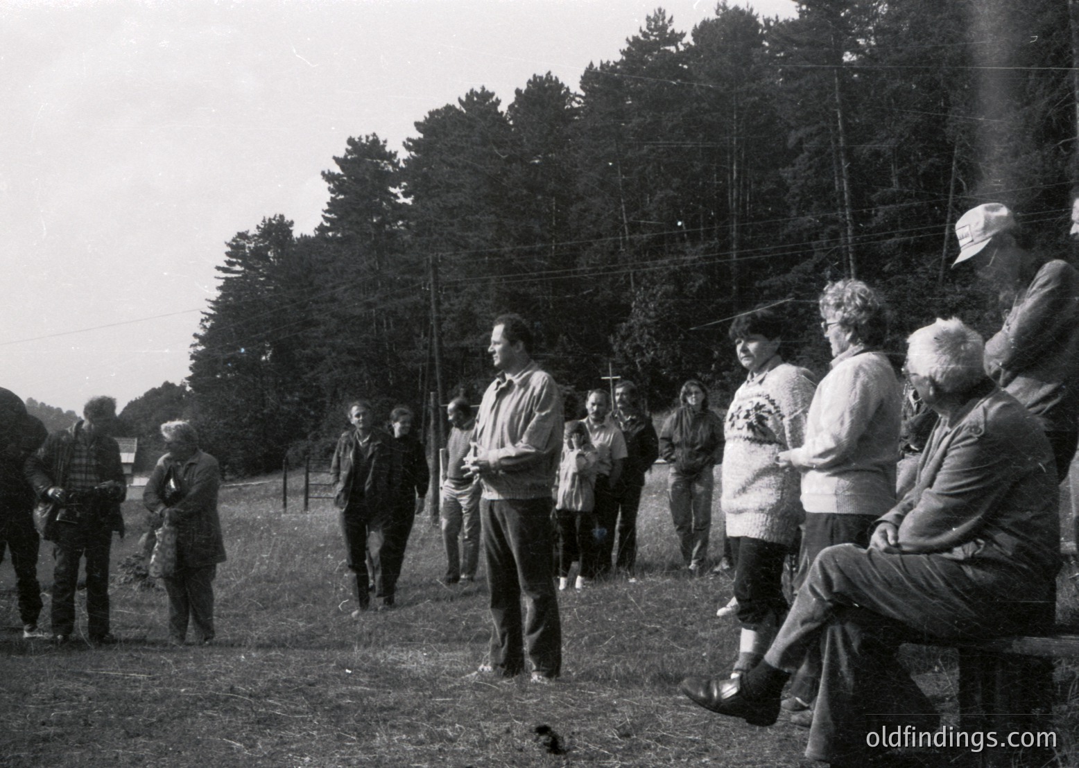 Group of adults in mid-20th-century outdoor gathering, likely 1950s–1960s. Central figure in dark jacket speaking to seated audience under tall trees. Casual attire suggests community event or lecture. Rural or park setting with open field and power lines visible.