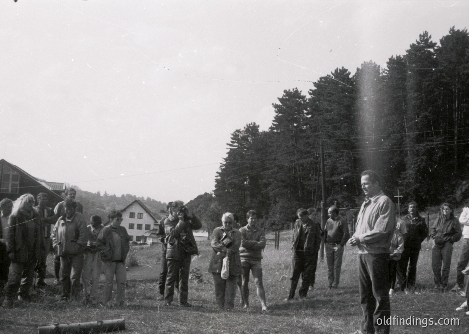 Mid-20th century group photo in rural setting, likely 1950s–1960s. Casual attire suggests informal gathering—men in trousers, women in dresses/blouses. Wooden farmhouse and dense forest backdrop indicate countryside. Sunlight filtering through trees creates lens flare.