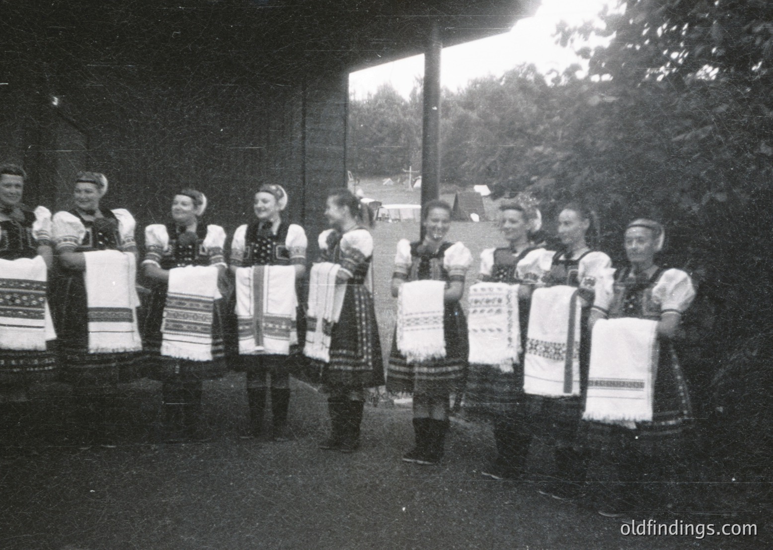 Group of women in traditional embroidered folk costumes, holding handkerchiefs with Cyrillic script, likely Bulgarian. Mid-20th century outdoor setting with greenery and wooden structure.