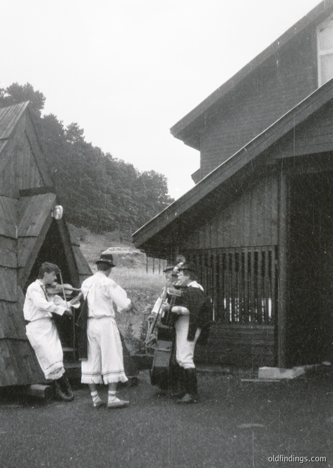Three musicians in traditional folk attire play outdoors near rustic wooden structures, likely a rural setting. Violinist in white embroidered vest, accordionist in dark vest with wide-brimmed hat, and another musician with a stringed instrument. Mid-20th century, Eastern European folk scene.