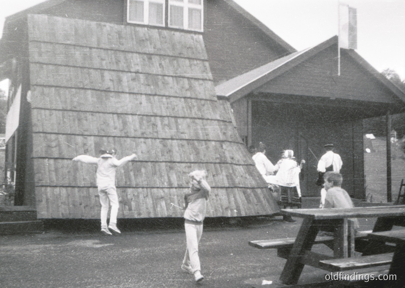 Black-and-white photo capturing a mid-20th-century outdoor scene. A man mid-jump against a wooden wall, while others observe from picnic tables. Wooden shingle architecture and casual summer attire ( )
