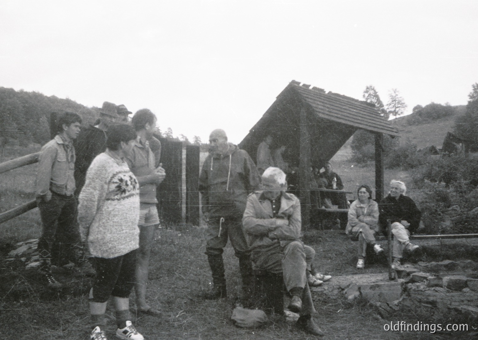 Group of 10+ individuals in a rustic outdoor setting, likely a hiking or camping area. Central figure seated on stone, surrounded by people in 1970s-era outdoor attire—flannel shirts, hiking boots, and caps. Wooden shelter with sloped roof in background. Overcast skies and forested hills suggest alpine or mountainous terrain.