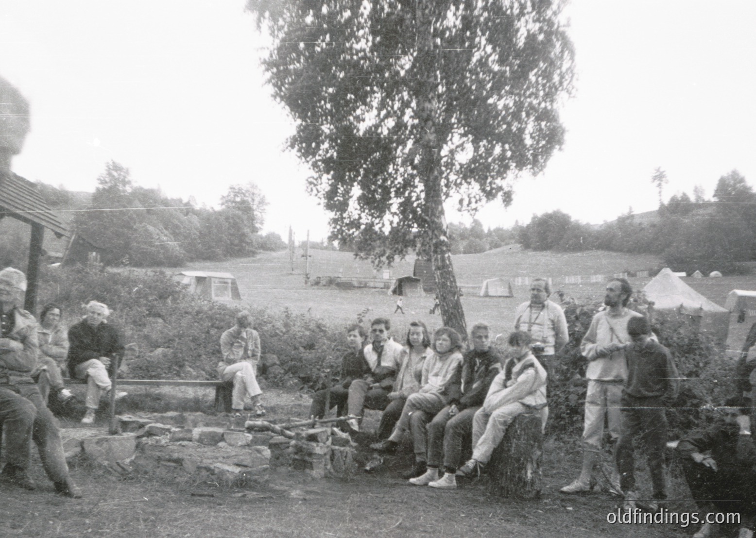 Group of 15+ individuals gathered around a campfire in a rural outdoor setting, likely 1960s–1970s. Wooden logs and makeshift seating suggest a casual, communal gathering. Tents and greenery in background indicate a camping trip or outdoor retreat.