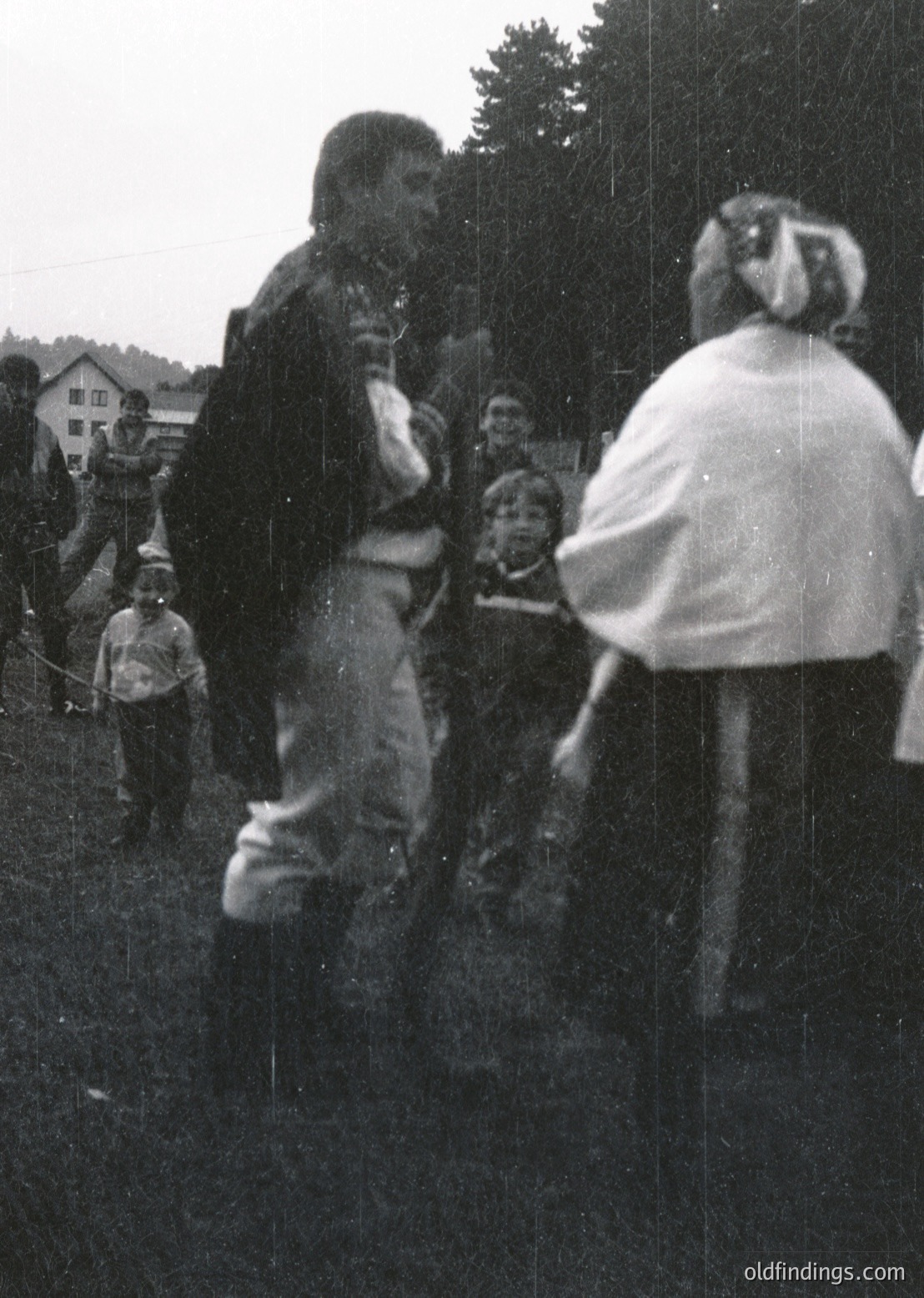 Vintage black-and-white photo of a rural outdoor gathering, likely a folk festival or harvest celebration. Central figures in traditional attire—one in a long coat and apron, another in a white shawl—engage in a ritualistic dance or game. Surrounding children and adults in casual 20th-century clothing observe. Wooden structures and greenery suggest a village setting, possibly Eastern Europe. Estimated 1950s–1960s.