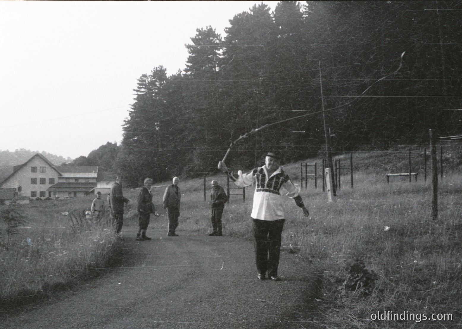 Black-and-white rural scene featuring a group of men playing frisbee on a grassy path, likely mid-20th century. Foreground man in apron/vest throws disc; others observe or walk. Background shows wooden farm buildings, dense trees, and utility poles. Evokes midcentury leisure and agricultural life.