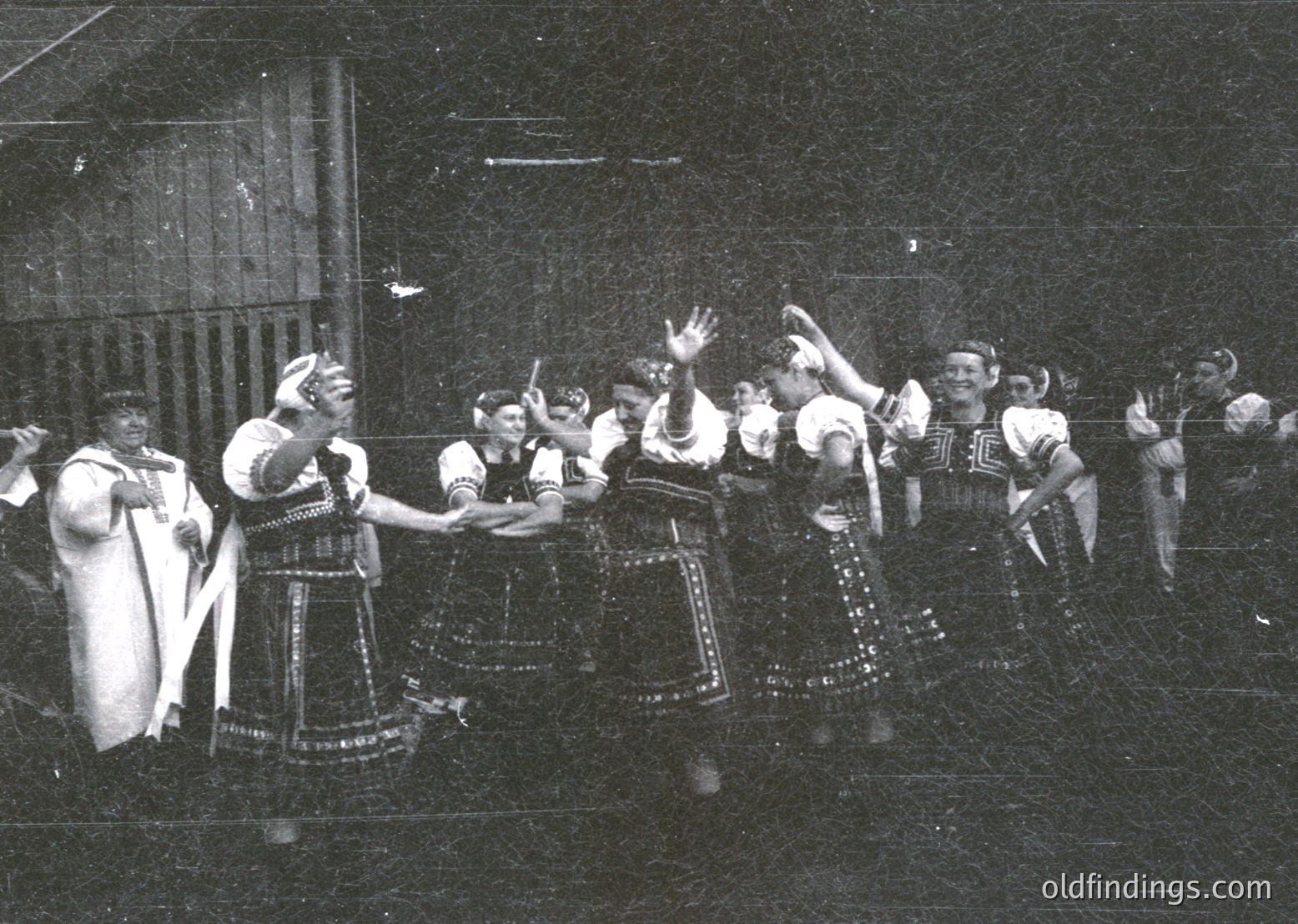Traditional folk dance performance in intricate embroidered costumes, likely Eastern European. Women in layered skirts with floral patterns and headscarves, men in vests and trousers. Stage lighting and blurred background suggest a cultural festival or folk festival setting, possibly 1950s–1970s.
