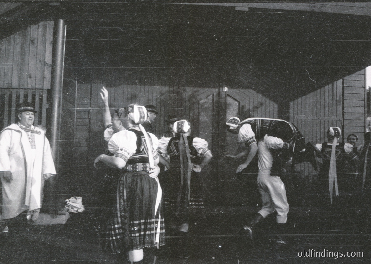 Black-and-white staged scene depicting traditional swordplay, likely a folk or theatrical performance. Central figures in period costumes—plaid skirt, sash, and headscarf—engage in mock combat with wooden swords. Spectators in formal attire observe. Setting appears to be an indoor hall or barn, suggested by wooden beams and rough walls. Style and attire hint at 19th–early 20th century European folk traditions.