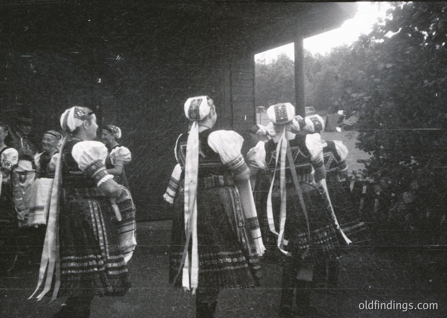 Traditional folk dancers in ornate embroidered costumes with wide belts and headscarves, performing indoors. Intricate patterns and layered skirts dominate their attire. Likely Eastern European cultural festival, mid-20th century.