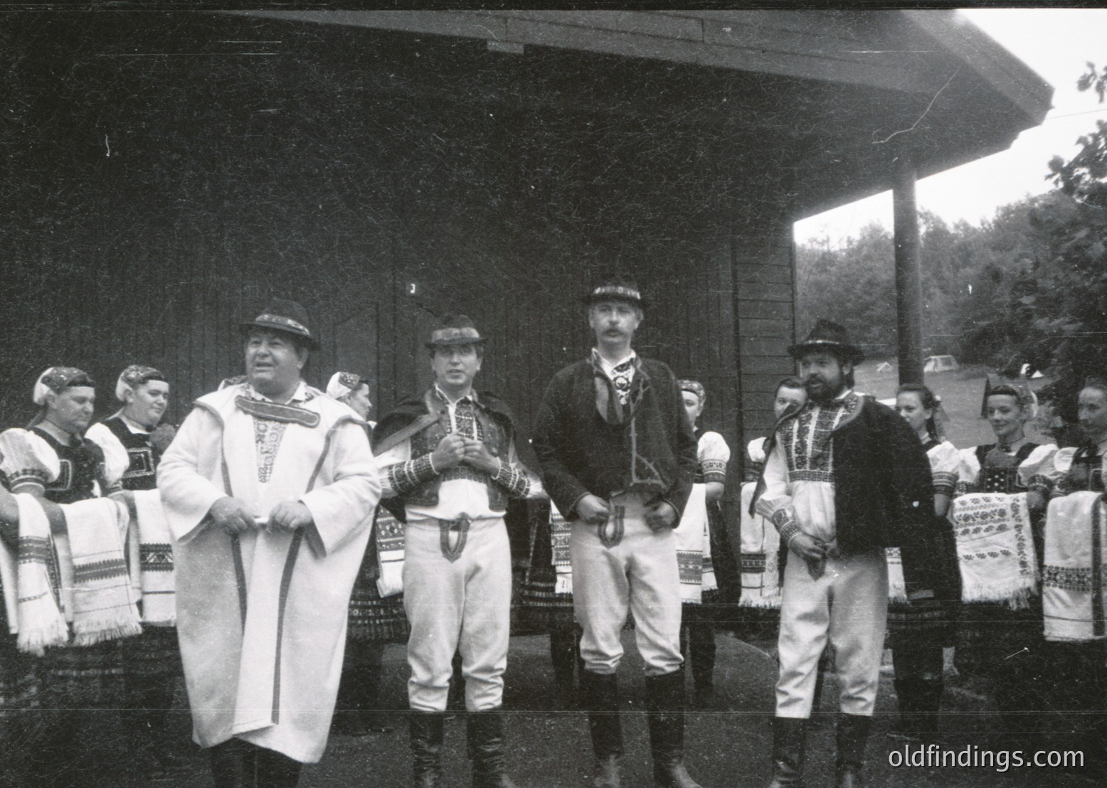 Black-and-white photo of a traditional folk ensemble performing outdoors, likely Eastern European. Men in embroidered vests, knee-length jackets, and wide-brimmed hats pose in a semi-circle. Decorative sashes and ornate belts accentuate their attire. Stage backdrop suggests a cultural festival or public event, possibly mid-20th century.