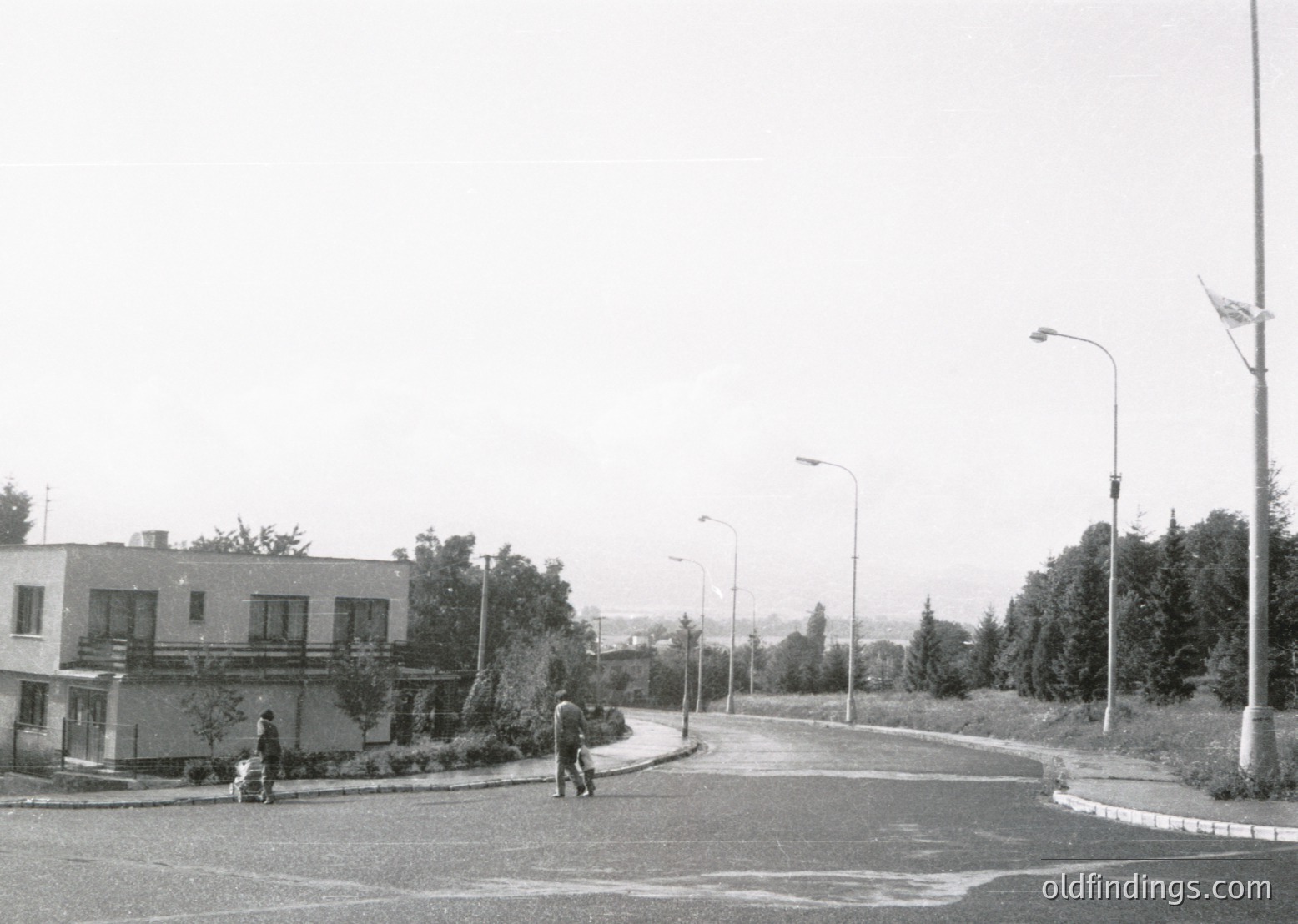 Mid-century urban street corner with minimalist Brutalist-style apartment blocks. Three pedestrians—one adult and two children—walk along a curved sidewalk. Overgrown greenery and lamp posts frame the scene. Likely Eastern Bloc architecture, 1960s–1970s.