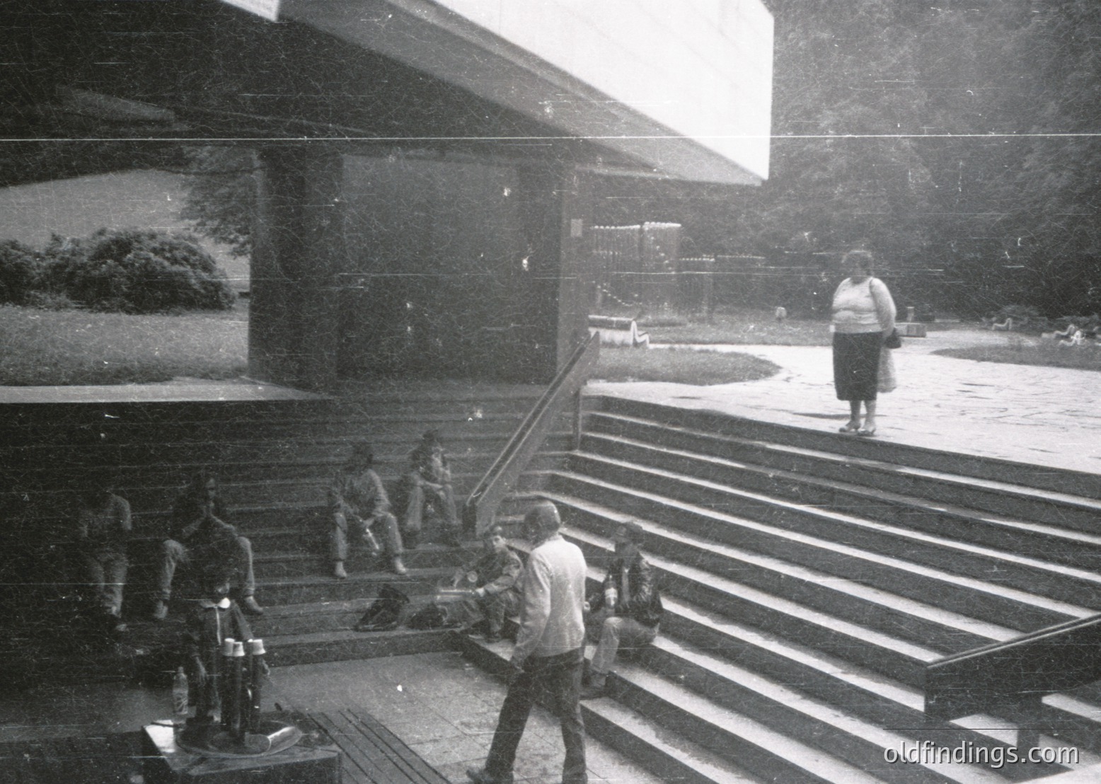 Vintage black-and-white photo of a grand, modernist stairwell with sweeping concrete steps. A man in mid-stride carries a long pole, while others descend or stand near bicycles. A lone woman walks away in the background, framed by a distant courtyard. Architectural details include geometric lighting fixtures and a large overhang. Likely Eastern Bloc-era institutional or public building, 1960s–1970s.