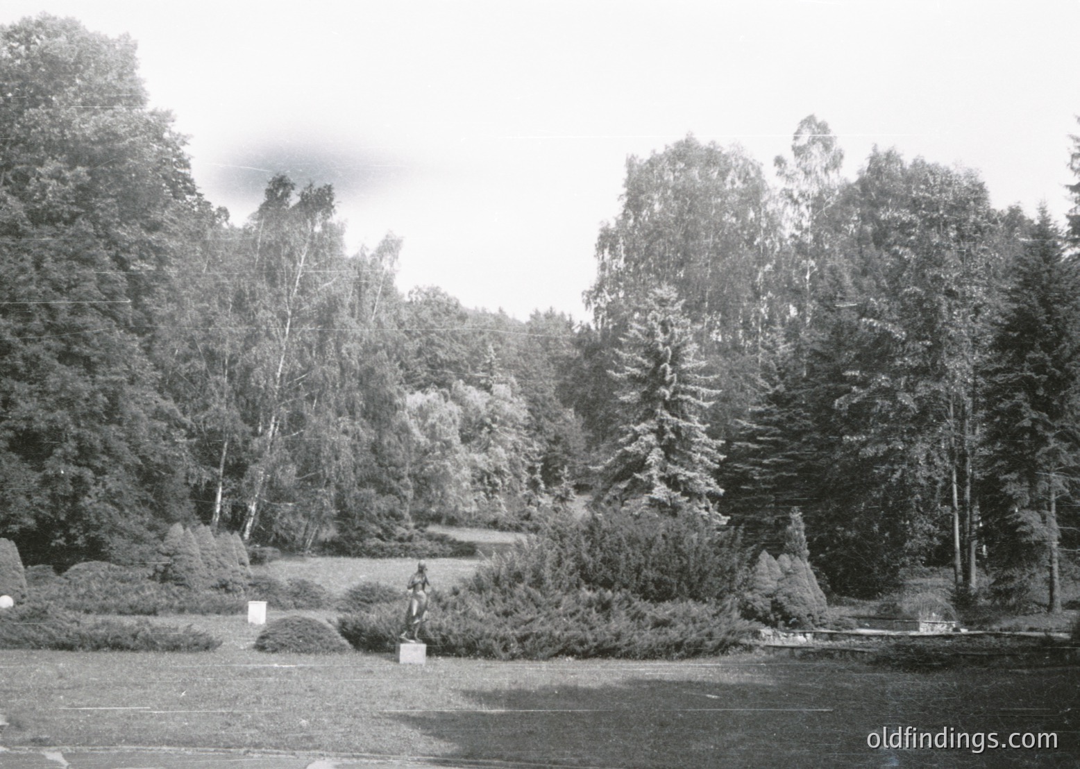 Black-and-white park scene featuring a statue of a seated figure on a stone pedestal, surrounded by manicured shrubs and trees. Pathway curves through landscaped greenery with scattered rocks. Mid-20th century design with classical elements.