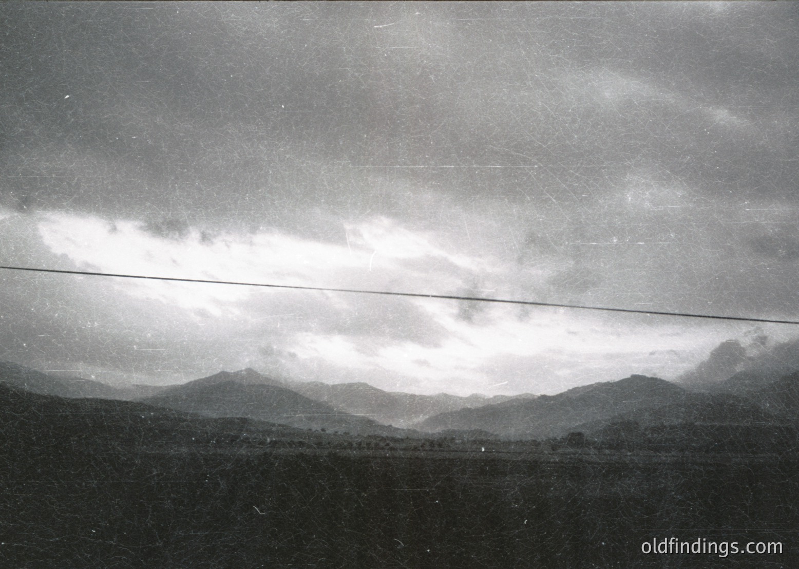 Vintage black-and-white landscape featuring low-hanging clouds over misty mountain ranges, with a horizontal power line cutting across the frame. The atmospheric haze suggests early morning or overcast conditions. Likely captured in a rural or alpine region during the mid-20th century.