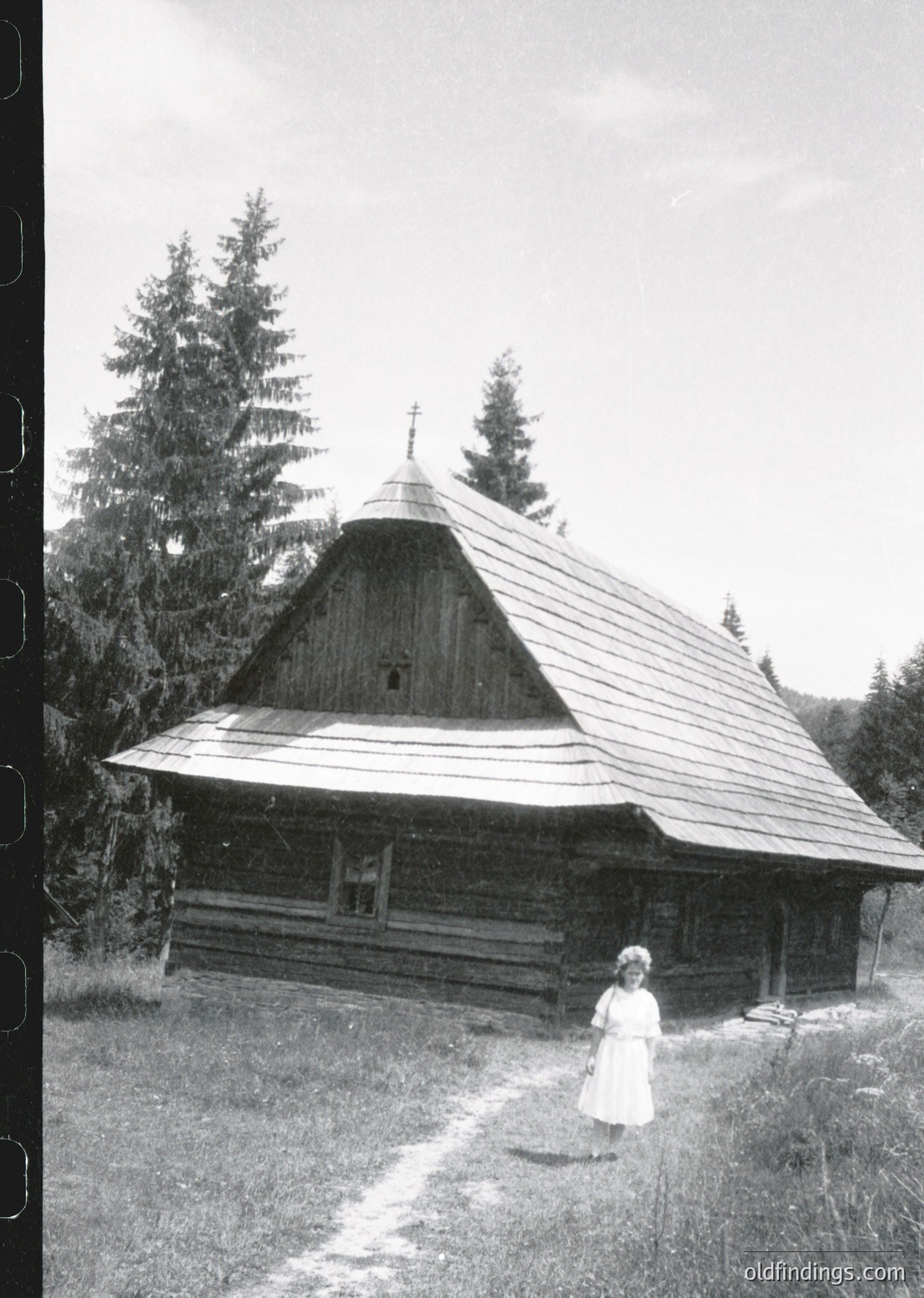 Traditional wooden church with steep gable roof and cross, surrounded by dense forest. Mid-20th century Eastern European rural architecture. Child in white dress poses near entrance.