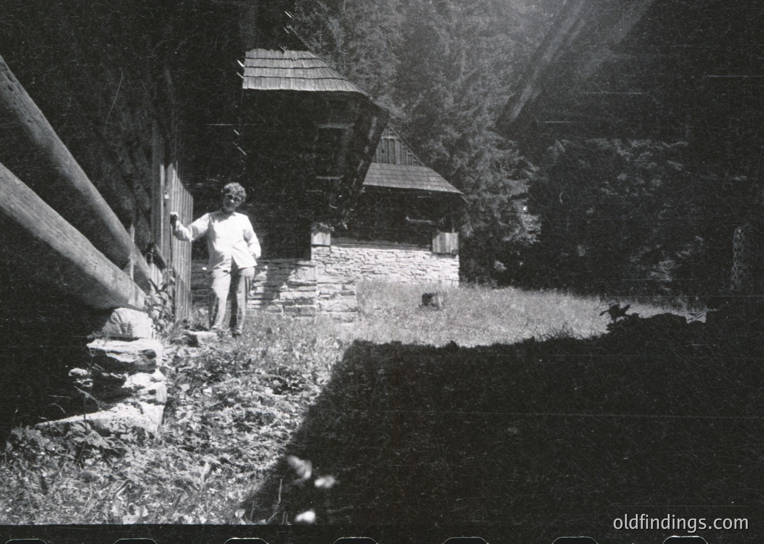 Rural stone cottage with wooden roofing, surrounded by dense forest. Two figures—one standing near a wooden gate, another seated on grass—suggest mid-20th century daily life. Sunlight filters through trees, casting dramatic shadows. Likely Eastern European countryside, 1950s-1960s.