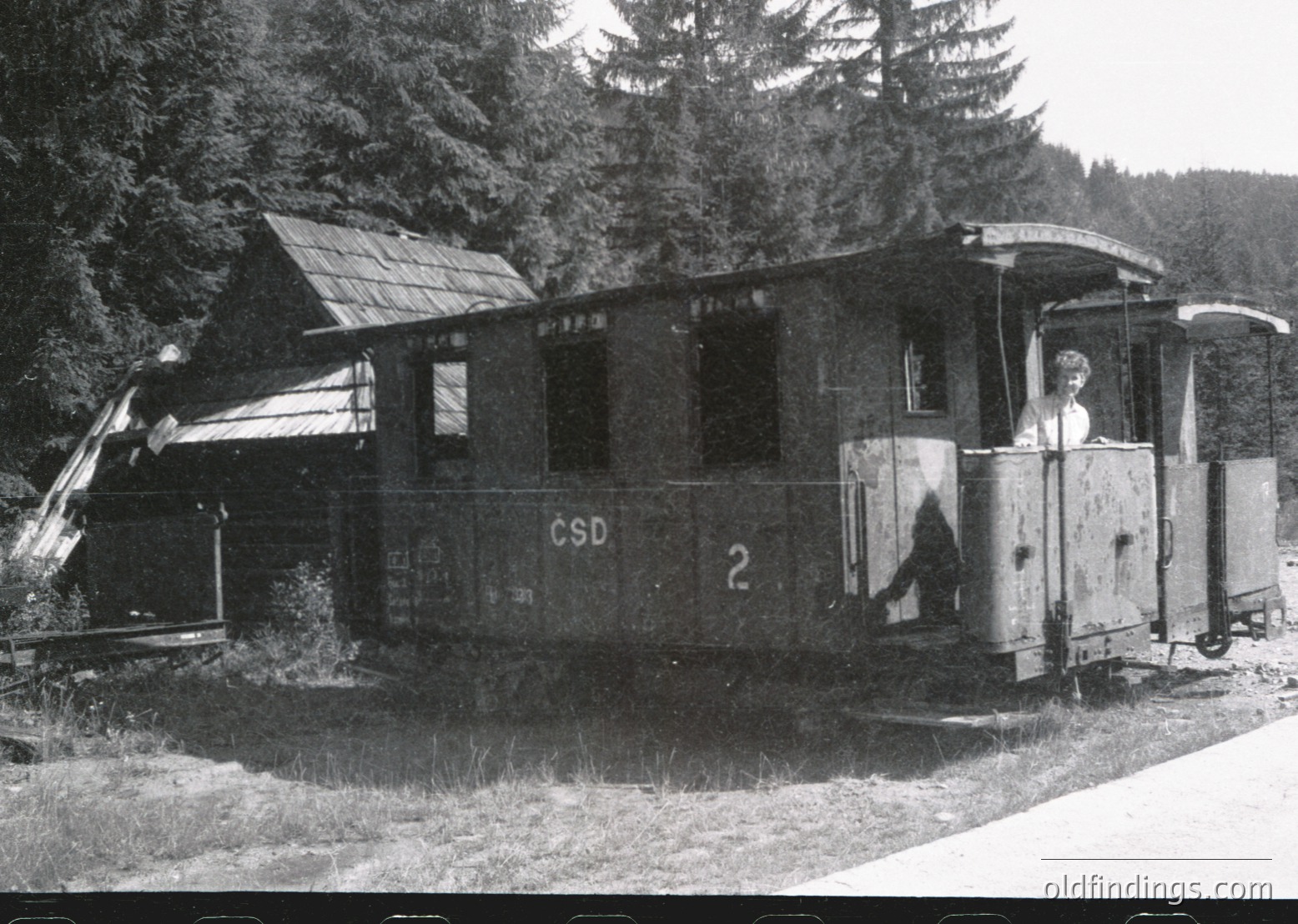 Vintage ČSD (Czechoslovak State Railways) narrow-gauge tourist train car, likely from the **1950s–1970s**, parked beside a rustic wooden station. Open door reveals interior seating; conductor stands at entrance. Surrounded by dense forest, suggesting a mountain or alpine route. Ideal for historical transport/railway archives. #ČSD