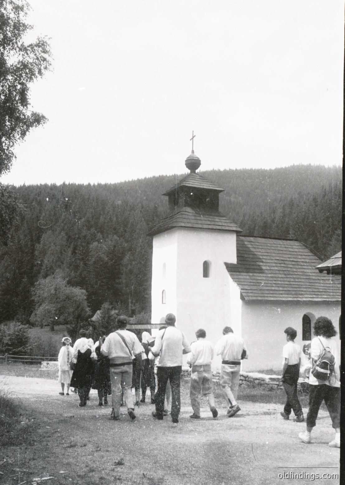 A group of people in casual 1970s attire—shorts, long-sleeve shirts, and backpacks—approaches a small, white Orthodox church with a bell tower and cross, set against a forested hillside. The scene suggests a rural or mountainous region, likely Eastern Europe. [Rural Orthodox church with bell tower surrounded by forested hills, 1970s-era group of visitors ]