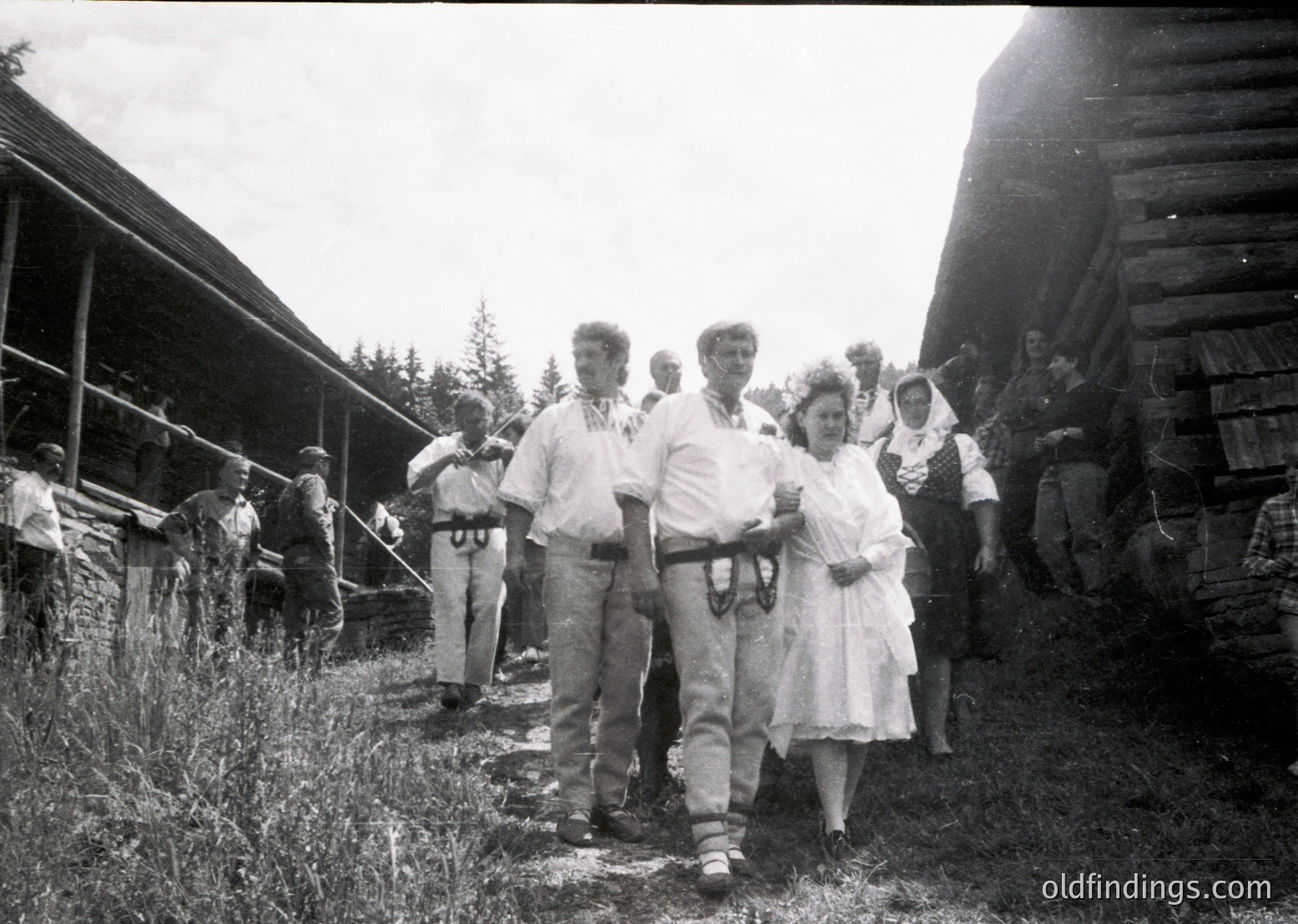 Rural gathering in mid-20th century, likely 1950s–1960s. Group of people in casual attire—men in trousers/suspenders, women in dresses—posing near rustic wooden structures and hay bales. Lush green field and overcast sky suggest agricultural setting.