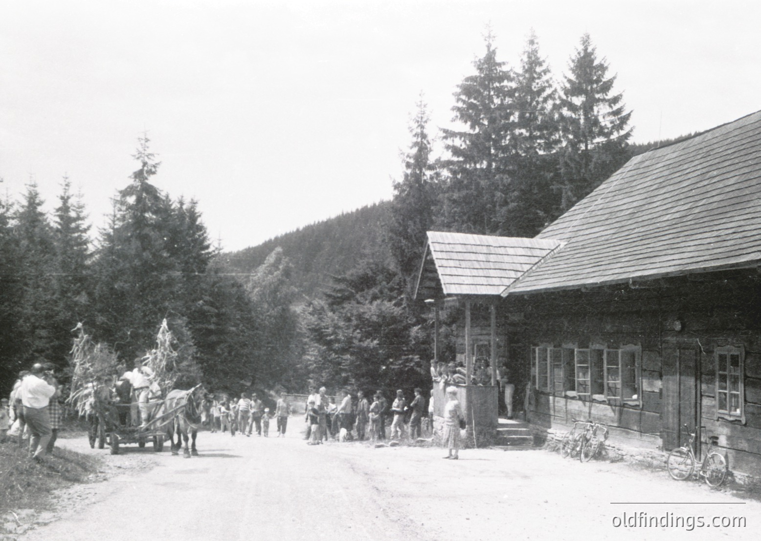 Black-and-white alpine village scene with wooden chalet-style buildings and dense pine forest. Crowd of early 20th-century mountain tourists, some with bicycles and horse-drawn carts, gathered near a rustic lodge entrance.