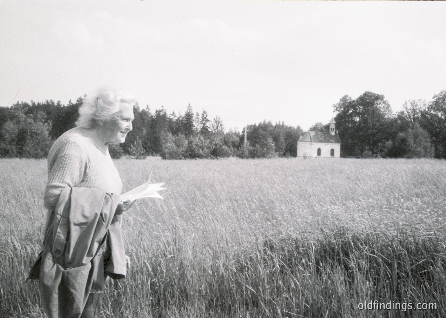 Black-and-white portrait of an elderly woman in a rural field, holding papers while wearing a sweater and a crossbody bag. Abandoned stone chapel in background among tall grass, surrounded by dense forest. Mid-20th century European countryside setting.