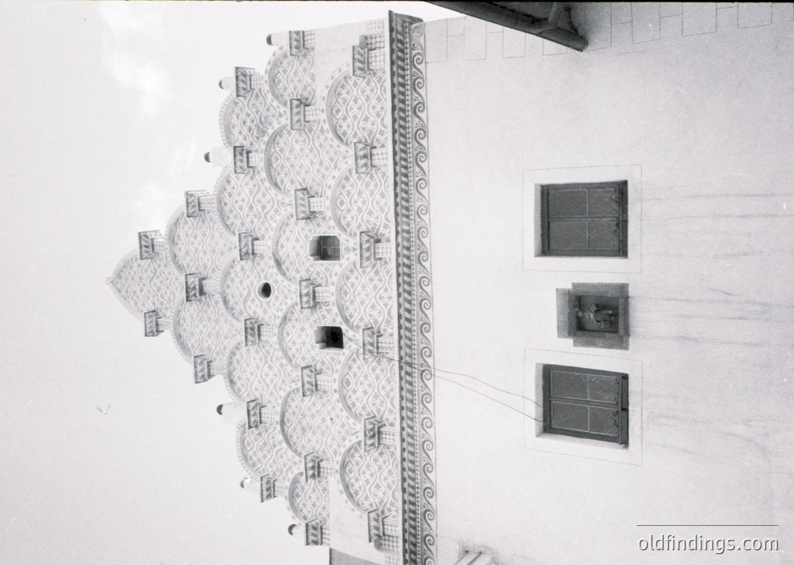 Mid-century Brutalist interior featuring geometric concrete ceiling with recessed circular alcoves. Ornate border with scrollwork frames the wall. Two small, barred windows on right reveal interior furnishings. Likely institutional or public building, 1960s–1970s.