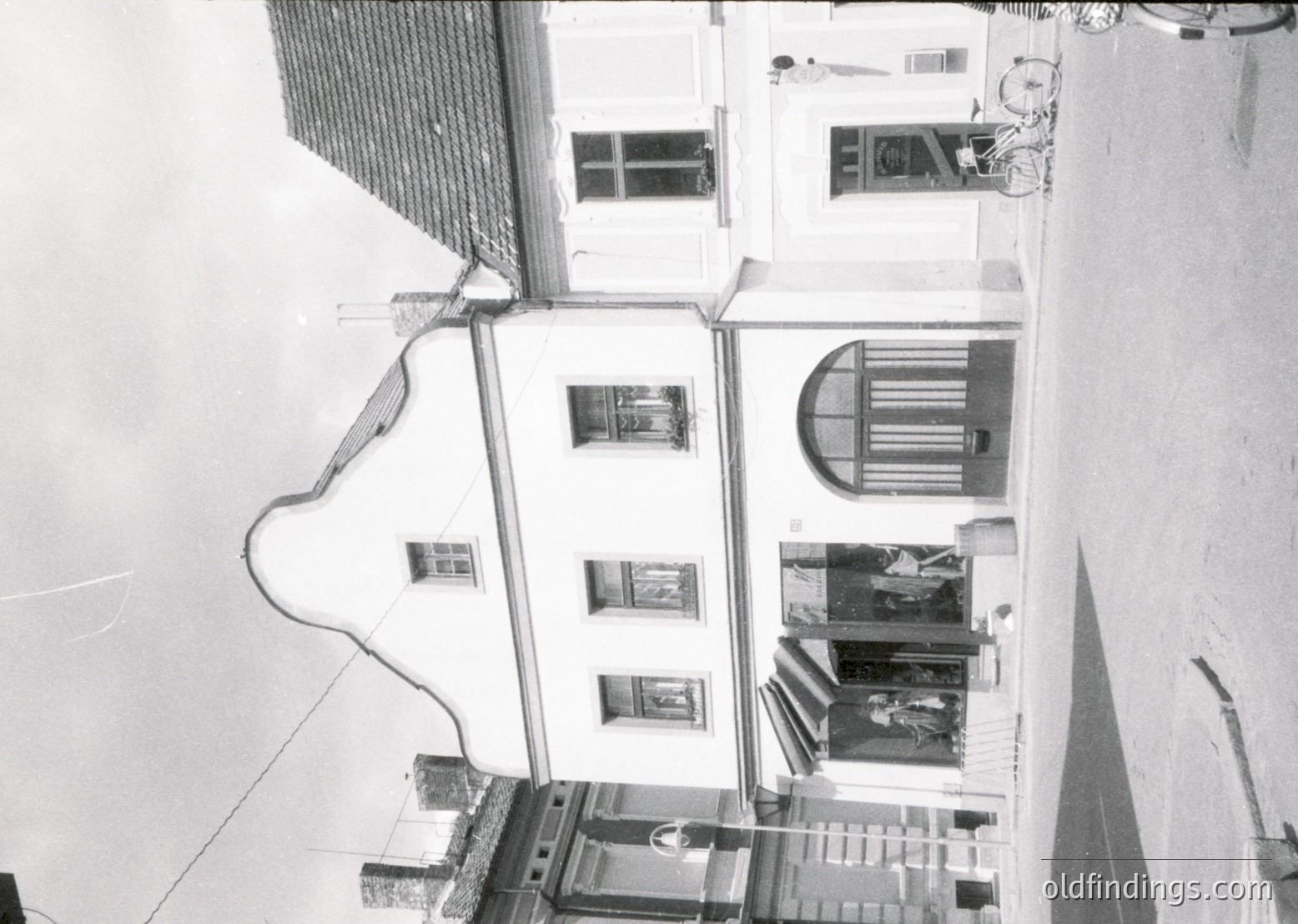 Mid-century urban courtyard with three-story residential blocks. Exposed pipes and wiring run along walls, indicating mid-20th-century construction. Balconies feature striped awnings and laundry, suggesting shared living spaces. Concrete staircases and tiled floors dominate the scene.