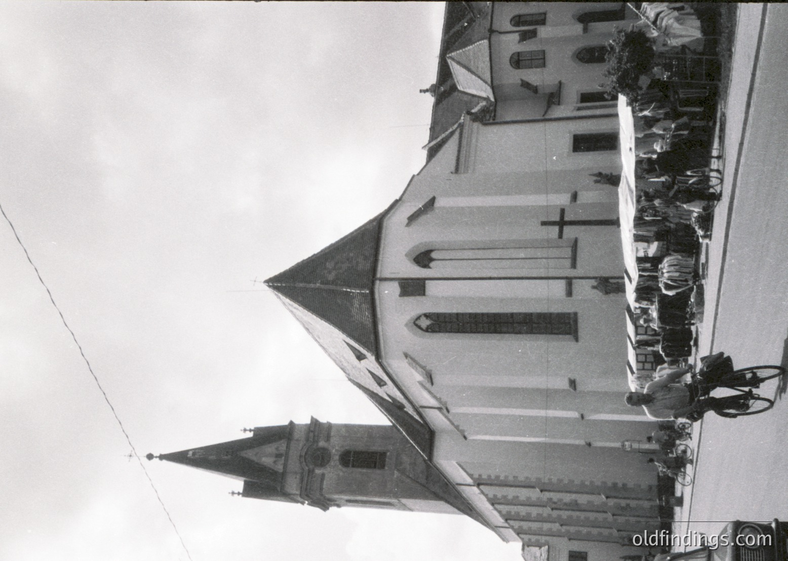 Historic church with steep gable roofs and pointed steeple, likely European Gothic Revival. Crowd of people gathered on rooftop terrace, suggesting a public event or ceremony. Low-angle perspective emphasizes architectural grandeur.