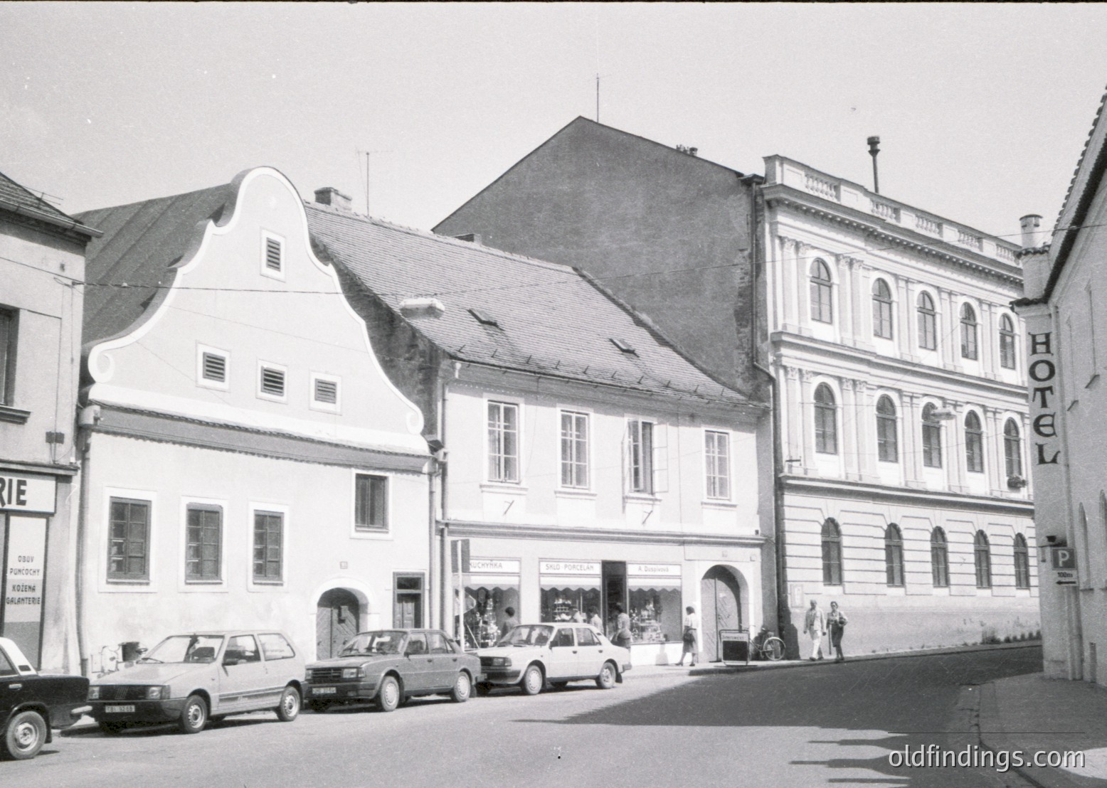 Mid-20th century European street scene featuring three-story buildings with classic European architecture: stepped gables, arched windows, and decorative cornices. The left building has a sign reading "Pizzeria" and a vintage car parked in front. The center building has a shop with large display windows. The right building is labeled "HOTEL" with a vertical sign. Empty street with minimal pedestrian activity.
