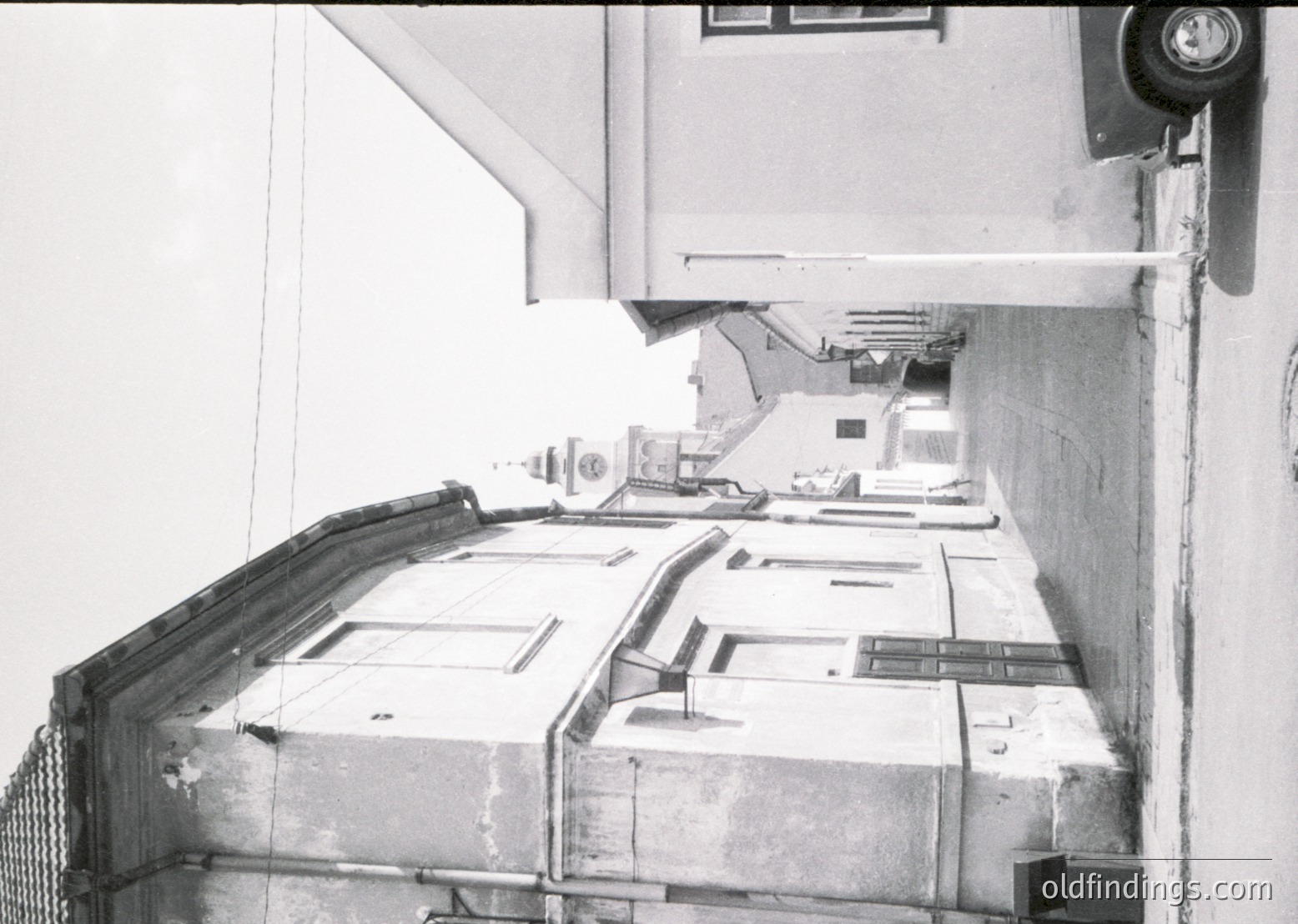 Industrial corridor with exposed concrete and metal railings, featuring a series of small, uniform windows aligned along the wall. Overhead, a pipe runs parallel to the floor. Likely a mid-20th century factory or warehouse setting.