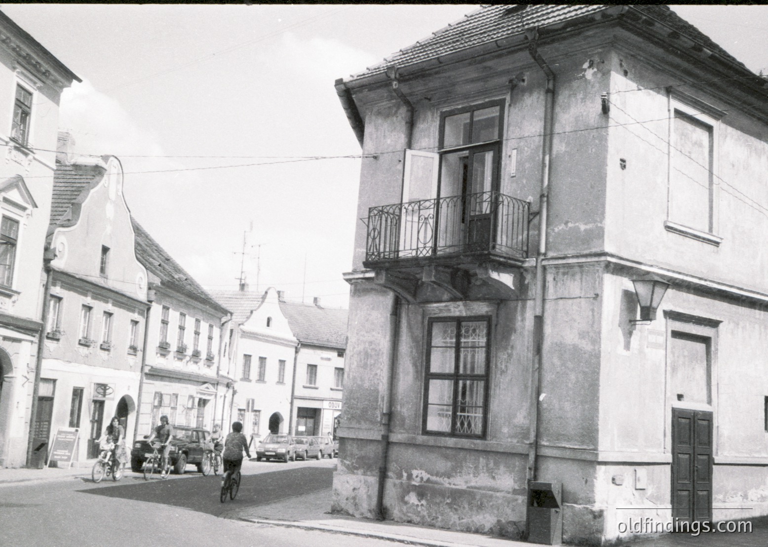 Mid-20th century European street scene featuring weathered two-story building with wrought-iron balcony and peeling plaster. Pedestrians, bicycles, and vintage storefronts suggest small-town daily life. Gabled roofs and cobblestone-like pavement hint at historic architecture.