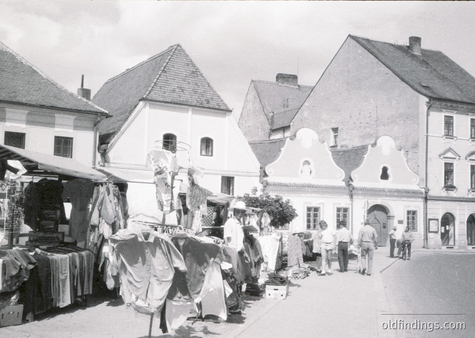 Vintage outdoor market featuring handmade textiles, pottery, and woven goods displayed on tables and racks. Traditional European architecture with steep gabled roofs and decorative facades dominates the scene. Crowd of casually dressed people browsing stalls, suggesting a community gathering. Likely late 20th century, possibly –.