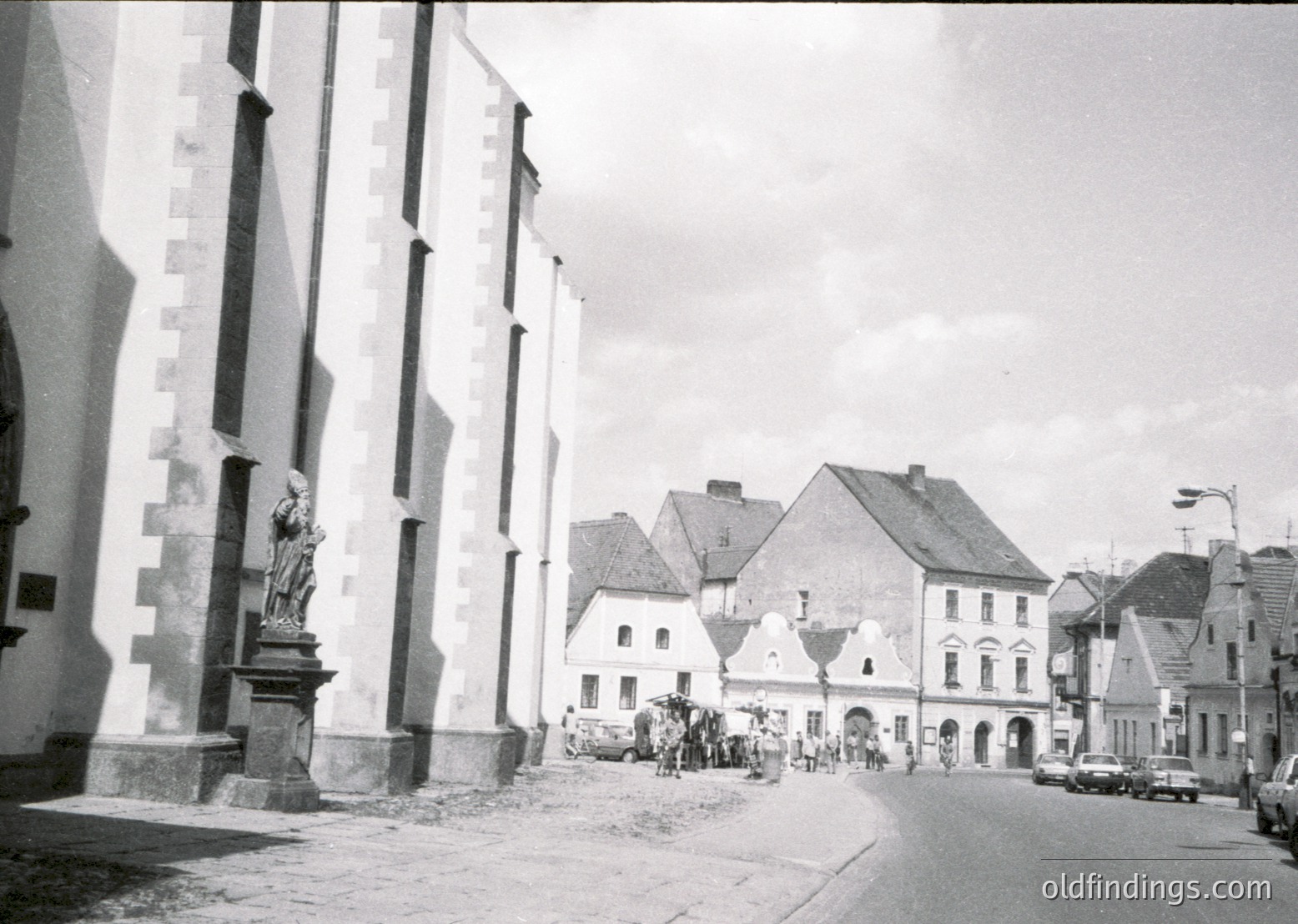 Mid-20th century European street scene featuring a stone statue of a robed figure on a pedestal beside a church façade with vertical striped columns. Pedestrians and vintage cars populate a cobblestone plaza with gabled buildings in the background. Likely Eastern Bloc architecture, 1950s–1960s.