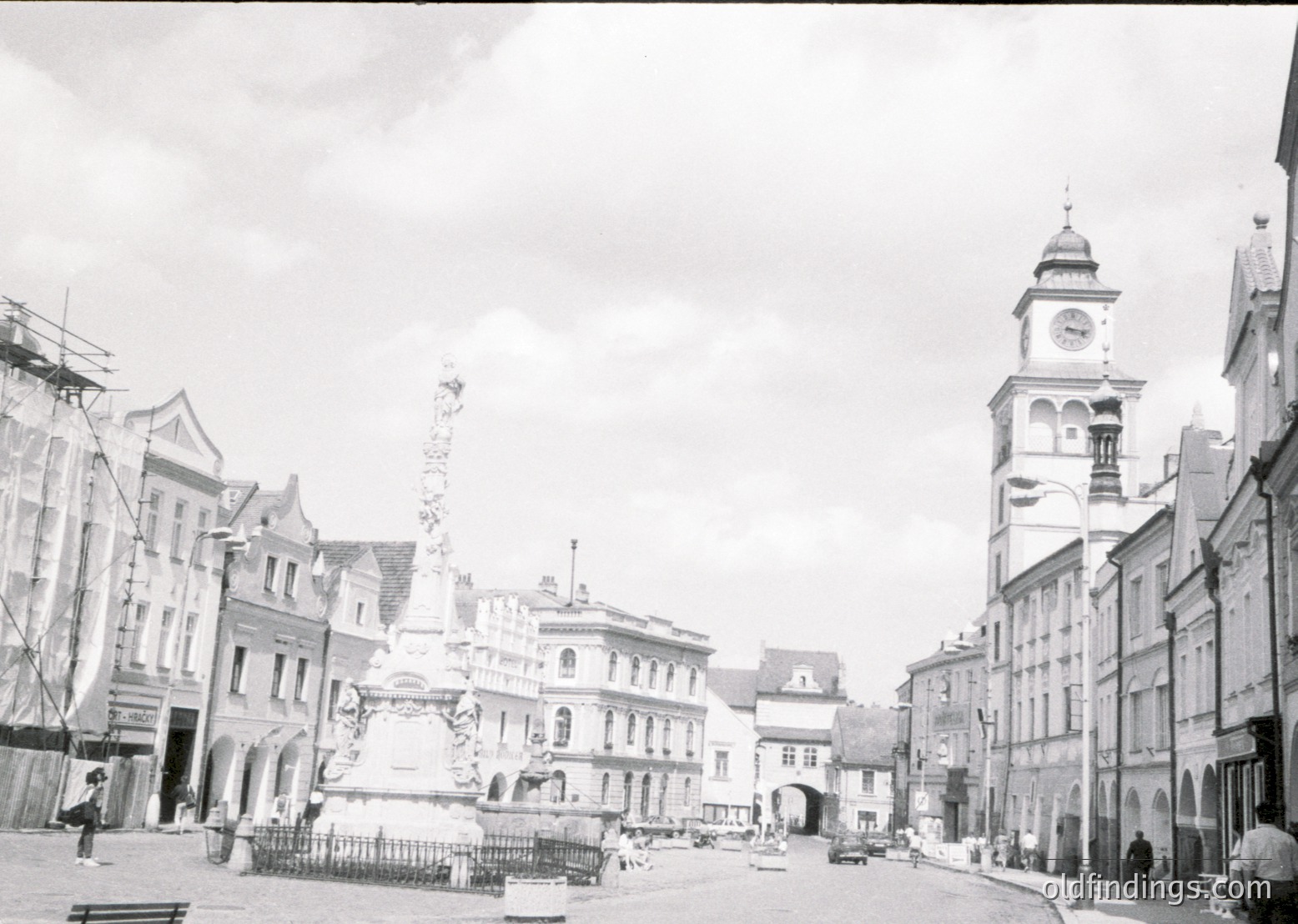 Black-and-white street scene featuring a central fountain with ornate sculptures and a tall column, flanked by historic European architecture. Clock tower with arched windows on right. Buildings exhibit 19th-century design elements like mansard roofs and decorative facades. Empty plaza suggests early-to-mid 20th century.