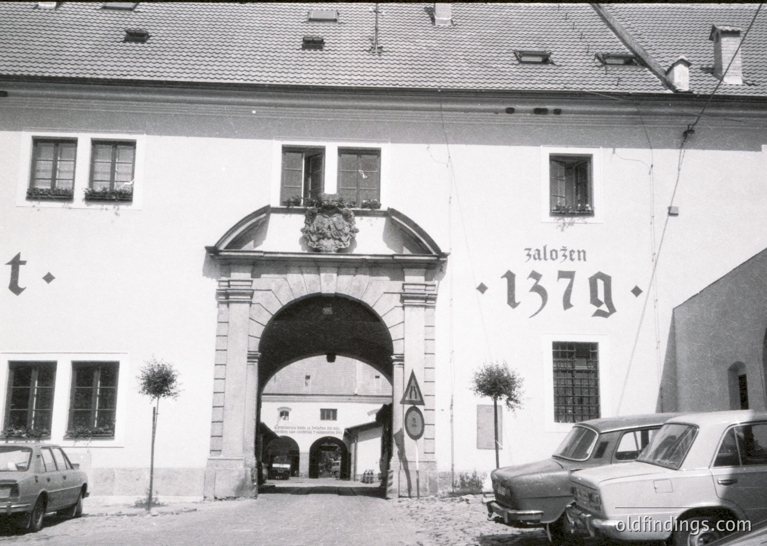 Historic stone archway entrance with "Zaloz̆en 1379" inscribed, indicating 1379 foundation. Classic European architectural details, including wrought-iron barred windows and small trees flanking the arch. Mid-20th century vintage cars parked nearby.
