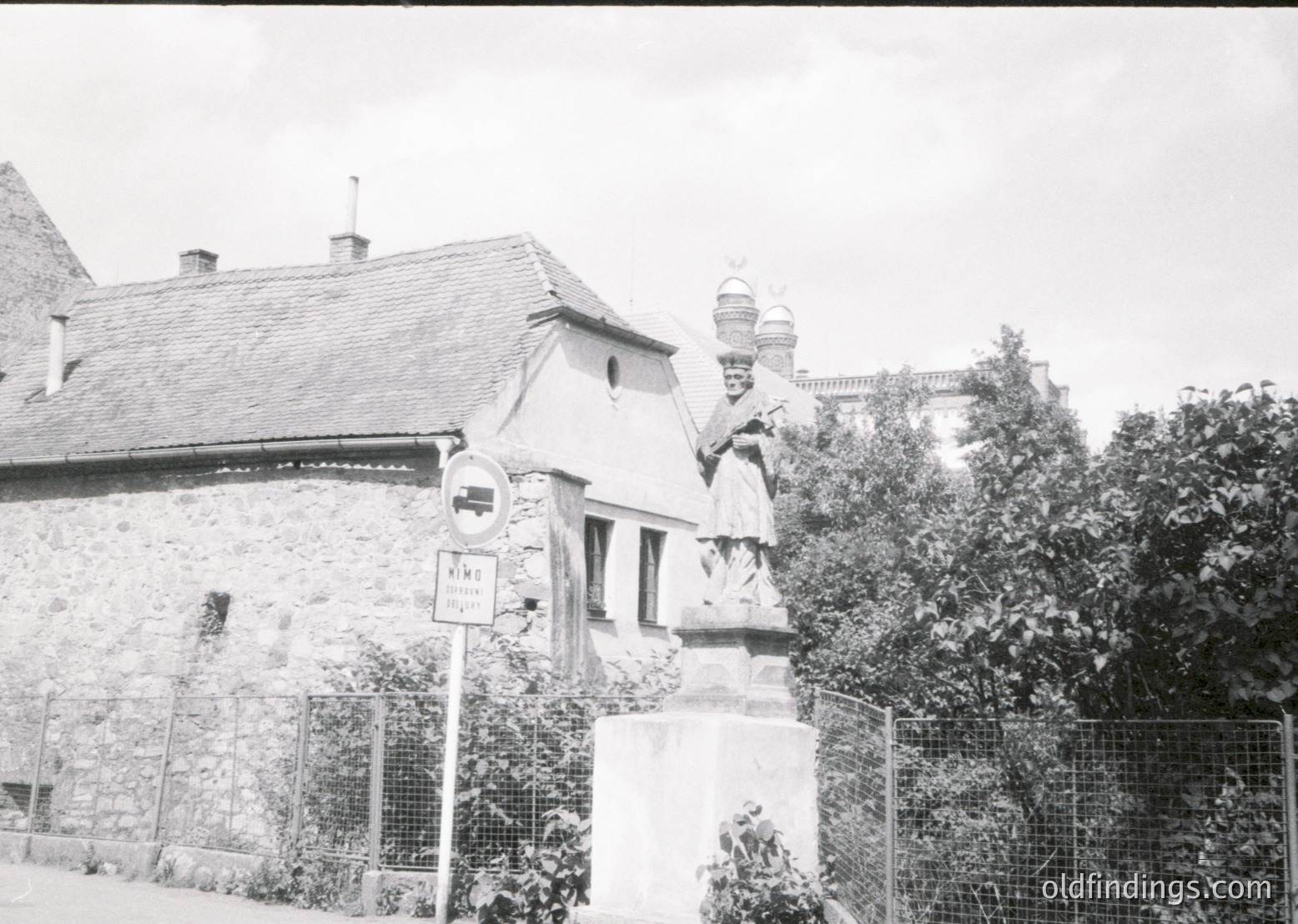 Black-and-white street scene featuring a stone statue of a robed figure holding a scroll, likely a historical or religious monument. Surrounding structures include a stone building with a pitched roof and chimney, and a domed tower in the background. A street sign reads "Ville" with "195" partially visible. Overgrown foliage and a chain-link fence frame the scene.