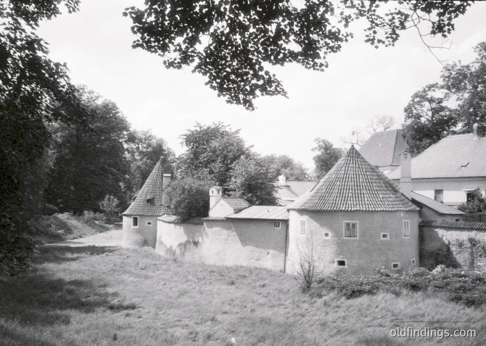 Historic rural estate featuring conical-roofed brick buildings with tiled roofs, surrounded by dense greenery. Likely European countryside architecture, mid-20th century.