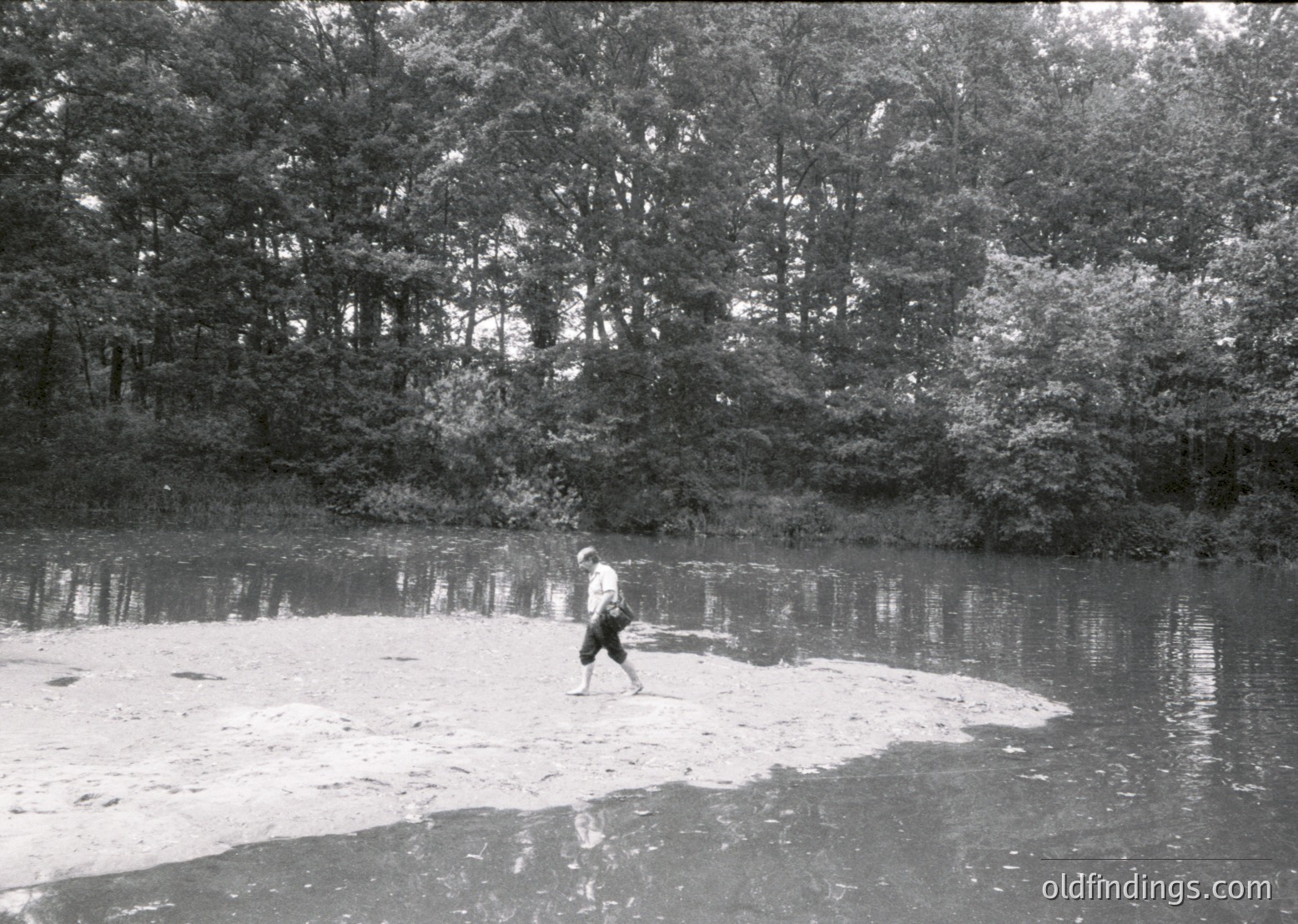 A lone individual wades through shallow, reflective water bordered by dense forest foliage. Black-and-white vintage photograph captures mid-20th century outdoor solitude.