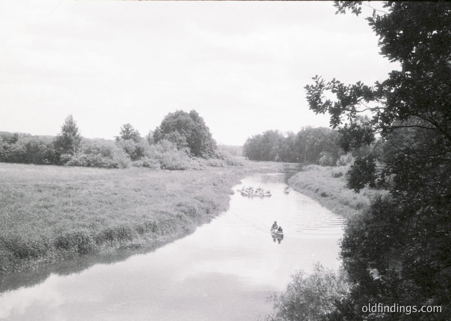 Vintage black-and-white scene of a narrow river flanked by dense reeds, with two small boats carrying people. One boat appears to be a rowboat; the other resembles a canoe or dinghy. Overcast skies and lush greenery suggest a rural, possibly agricultural setting. Style and lighting indicate mid-20th century (1940s–1960s).
